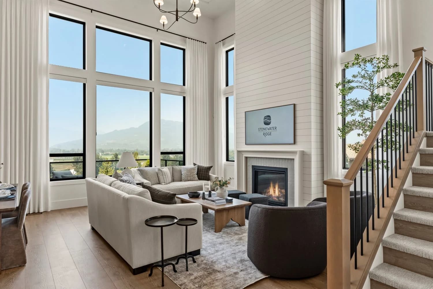 Stonewater Ridge – A bright living room features soaring double-height ceilings, a white shiplap fireplace with a mounted TV, and expansive floor-to-ceiling windows offering scenic mountain and valley views. The space is furnished with neutral sofas, dark armchairs, light wood floors, and a carpeted staircase along the right.