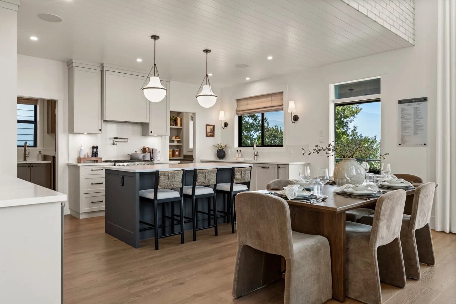 Stonewater Ridge – A bright, open-concept kitchen and dining area featuring light hardwood floors, white tongue-and-groove ceiling paneling, and a dark grey kitchen island with barstool seating beneath hanging pendant lights. A wooden dining table with upholstered beige chairs is set in the foreground, with large windows and a glass door in the background overlooking lush green trees under daylight.