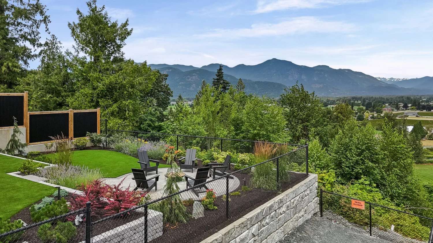Stonewater Ridge – A terraced outdoor patio and lawn area features dark Adirondack chairs arranged in a circle, surrounded by landscaped gardens, lush trees, and chain-link fencing atop a stone retaining wall. Beyond the patio, an expansive valley unfolds toward a majestic mountain range beneath a light blue, partly cloudy daytime sky.
