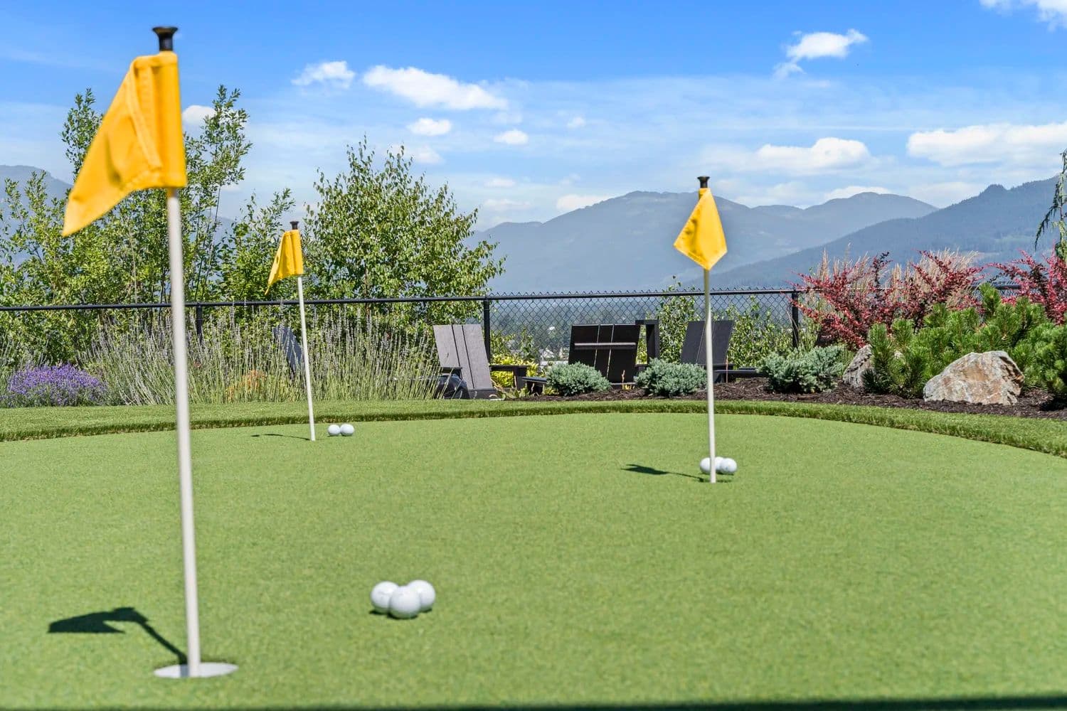 Stonewater Ridge – An outdoor putting green amenity features bright yellow flags, golf balls, and well-manicured artificial turf under bright daylight. In the background, landscaped gardens with Adirondack chairs overlook a scenic mountain range beneath a clear blue sky.