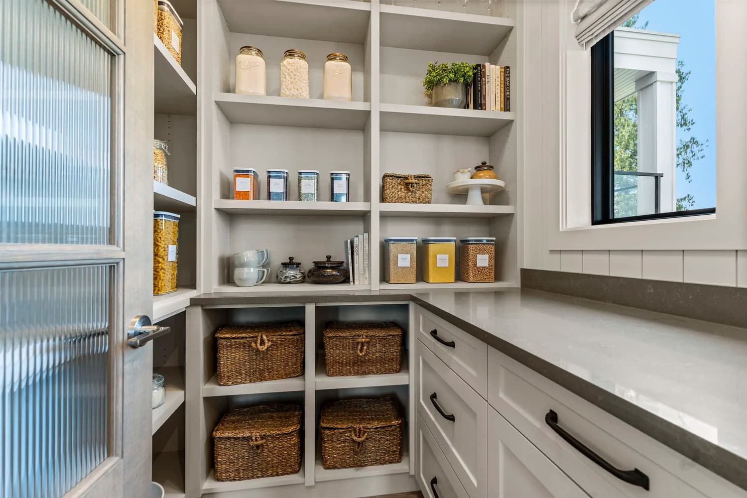 Stonewater Ridge – A well-organized walk-in pantry featuring custom white open shelving and lower cabinetry topped with gray quartz countertops. Clear storage canisters, woven wicker baskets, and cookbooks are neatly arranged on the shelves, with natural daylight entering from an adjacent window.