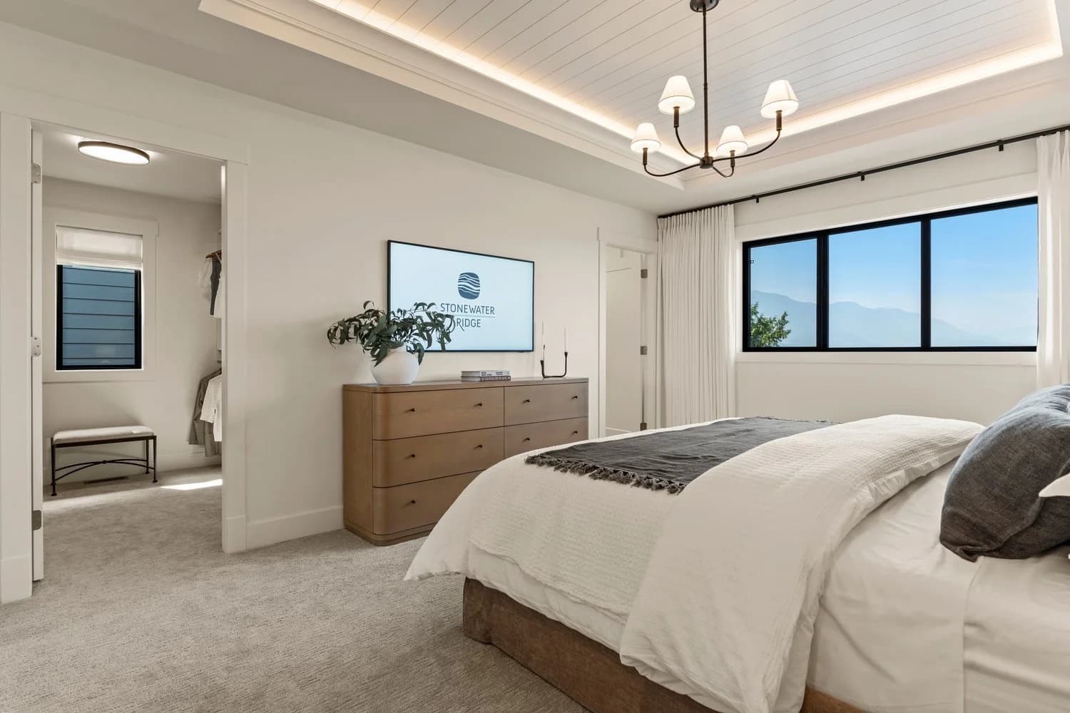 Stonewater Ridge – A bright modern bedroom features a tray ceiling with cove lighting, shiplap detailing, and a classic chandelier above a neatly made bed. Opposite the bed sits a light wood dresser with a wall-mounted TV, adjacent to a wide window framing distant mountain views and an open doorway leading to a walk-in closet.