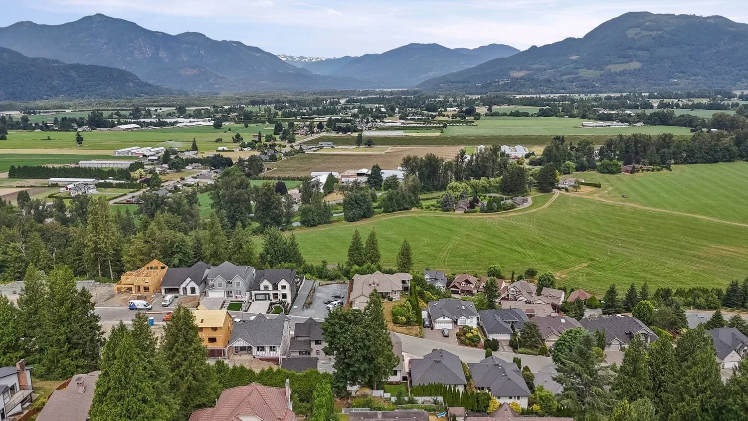 Stonewater Ridge – An elevated shot overlooks a suburban residential neighborhood with homes under construction, backing onto expansive green agricultural fields and a wide rural valley. In the background, prominent mountain ranges with lush forested slopes sit under a slightly overcast daytime sky.