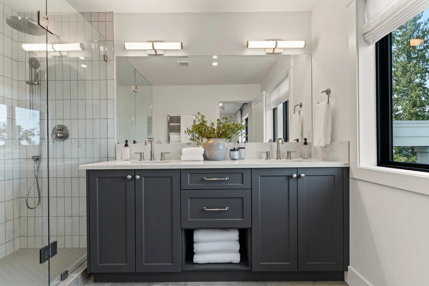 Stonewater Ridge – A modern bathroom features a dark charcoal double vanity with a white countertop, brushed metal hardware, and a large mirror topped by two horizontal light fixtures. To the left is a glass-enclosed walk-in shower with vertical white subway tiles and a rain showerhead, set against bright white walls with a window looking out toward green trees.