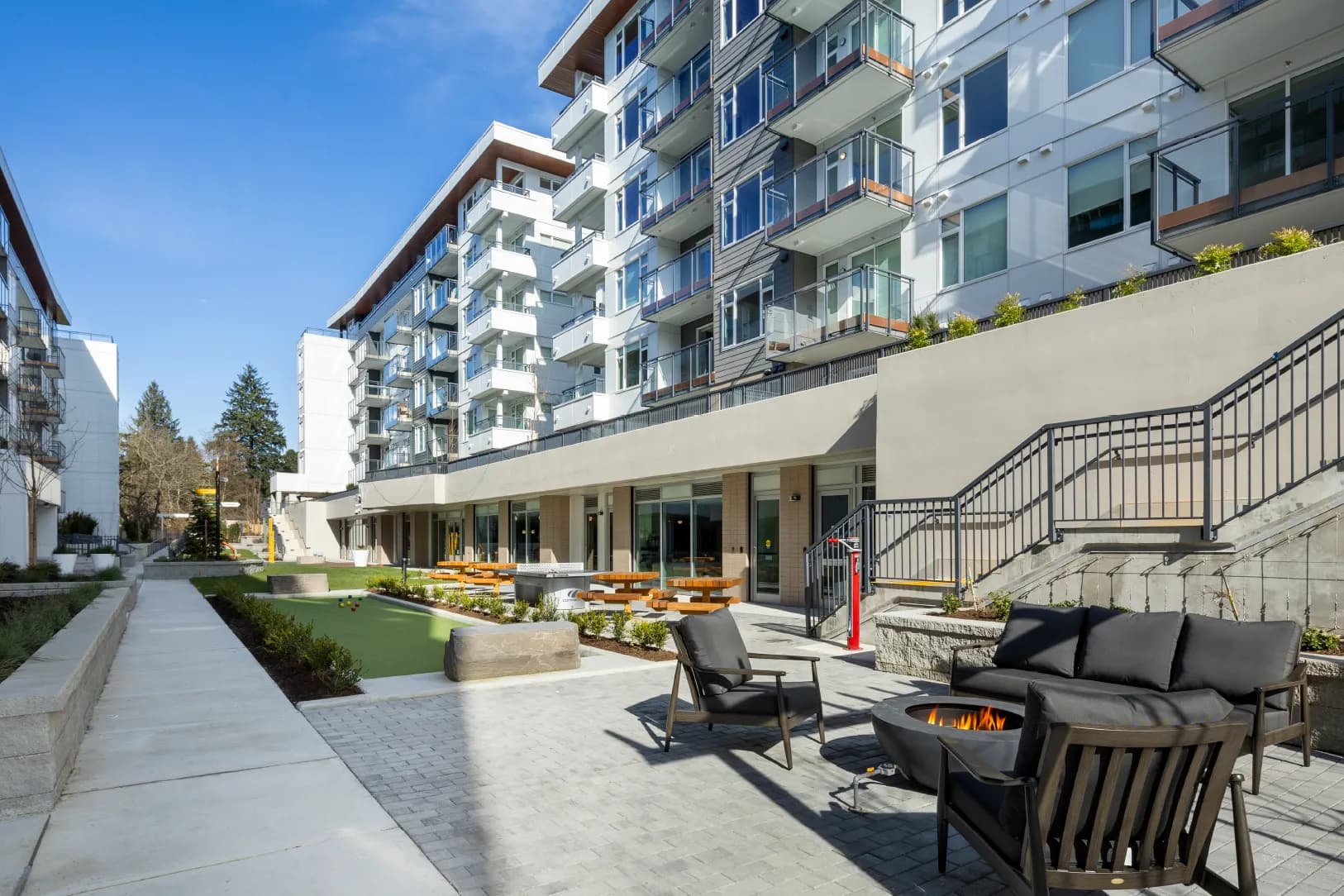 Hartley – An outdoor community courtyard nestled beside modern multi-story residential buildings, featuring a paved lounge area with dark cushioned armchairs and a round fire pit in the foreground. In the background, the landscaped terrace includes a green bocce ball or putting turf, picnic tables, and concrete retaining walls under bright blue daytime skies.