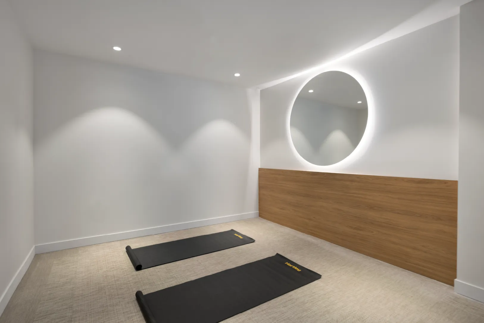 Hartley – A minimalist yoga or fitness studio features two black exercise mats laid out across a textured light-colored floor. Crisp white walls are accented by warm wood paneling, recessed ceiling spotlights, and a large circular mirror with backlight illumination.