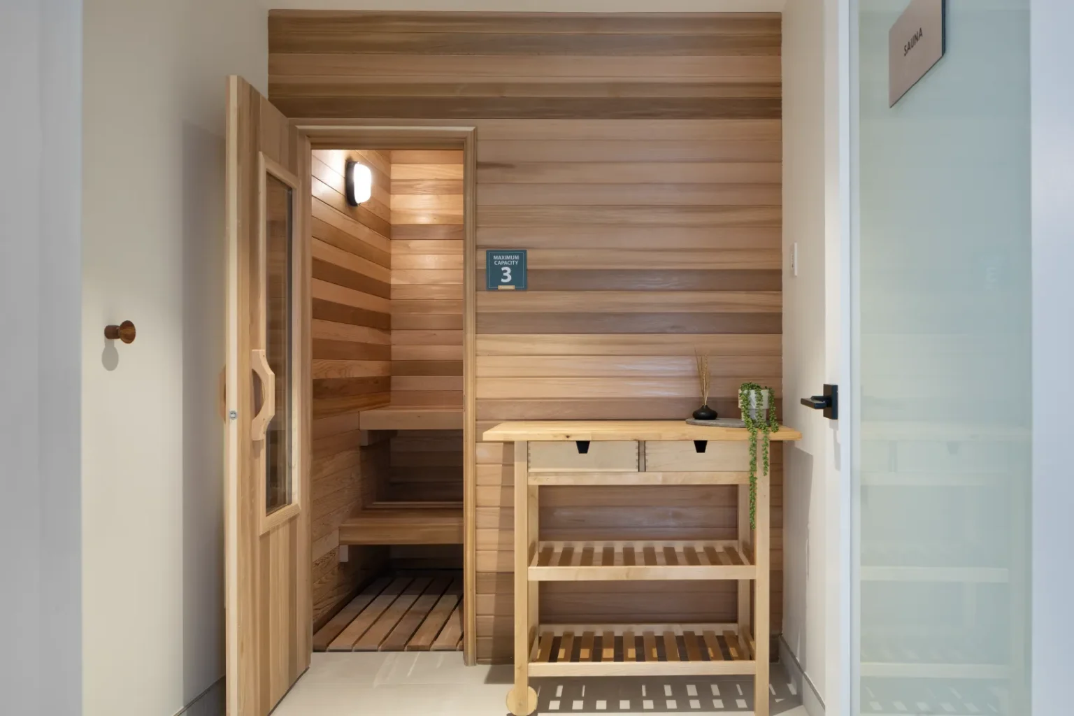 Hartley – A modern indoor sauna area features an open wooden door revealing tiered cedar benches and warm interior lighting. Outside the sauna, a matching wood-paneled accent wall is complemented by a light wood utility cart holding a small plant and diffuser, creating a calm spa atmosphere.