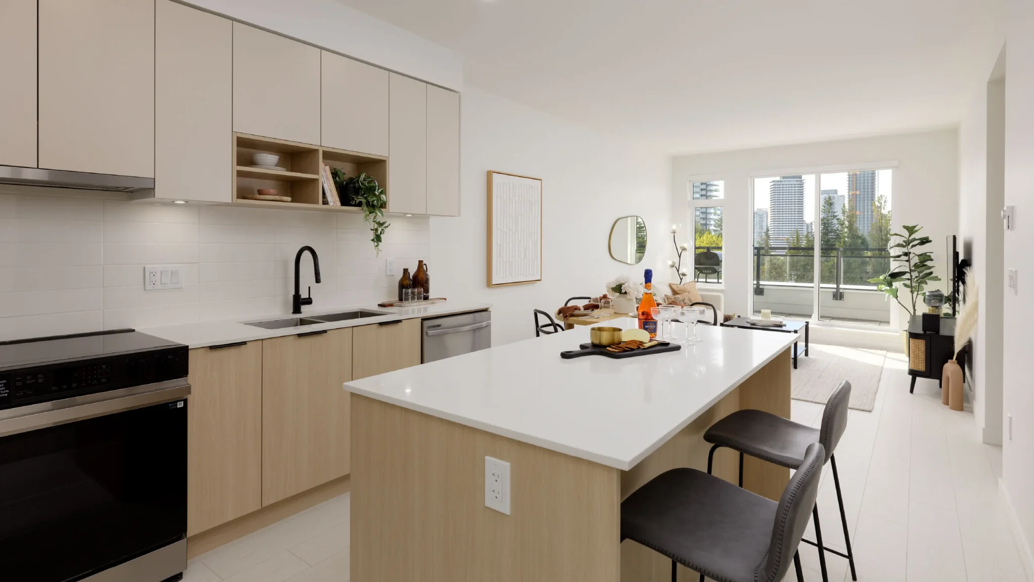 Hartley – A bright, open-concept kitchen and living area featuring a large white quartz island with barstools, light wood and beige cabinetry, and stainless steel appliances with matte black fixtures. Beyond the dining space and living room, large sliding glass doors open onto a balcony overlooking treetops and modern high-rise towers in the daylight.