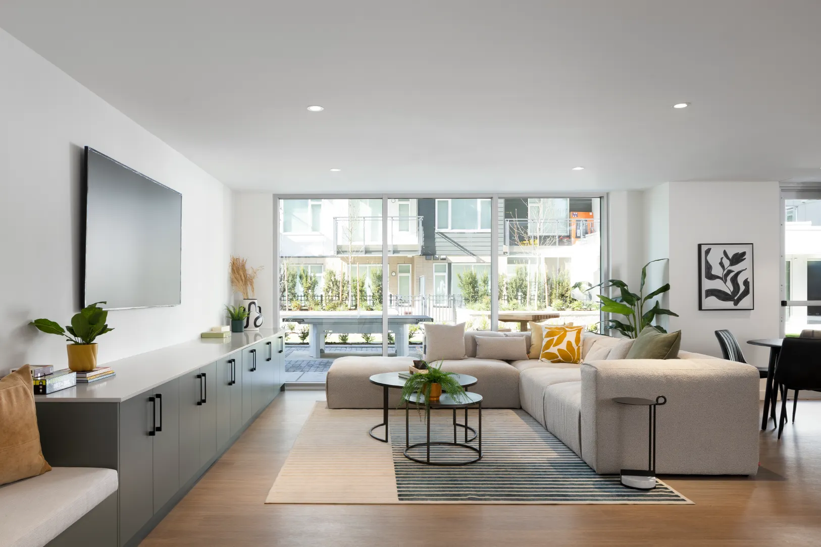 Hartley – A bright, modern indoor lounge furnished with a large light gray sectional sofa, round coffee tables, and a wall-mounted flat-screen TV above built-in gray cabinetry. Floor-to-ceiling glass doors open directly to an outdoor patio featuring an outdoor ping-pong table and views of neighboring residential townhomes in daylight.