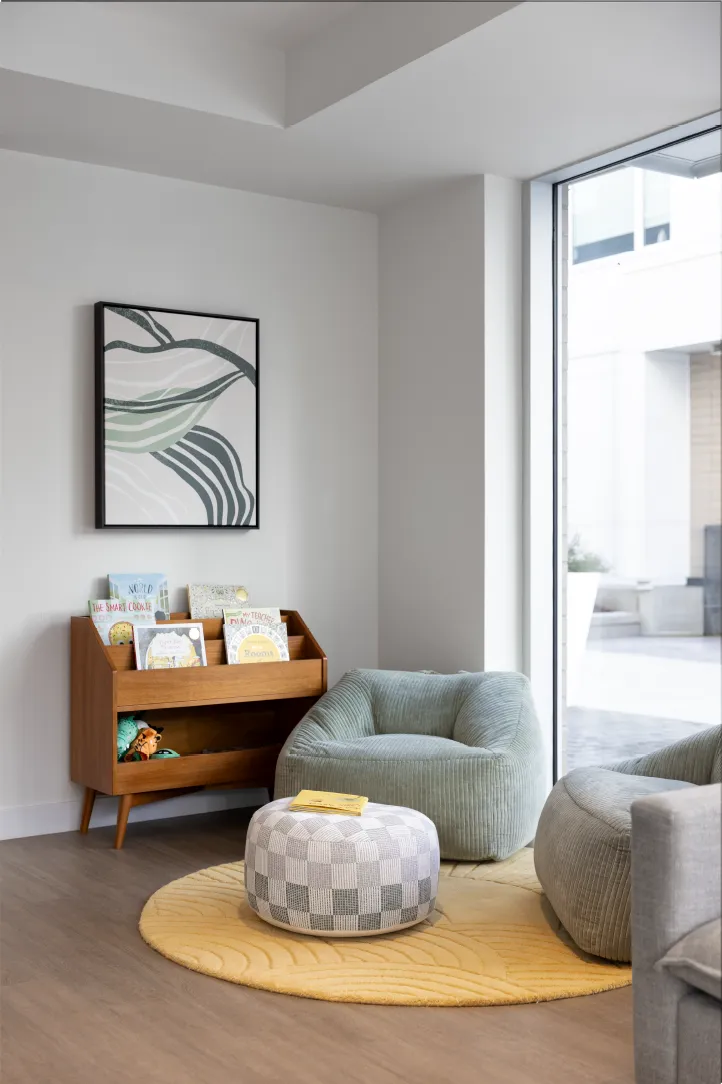 Hartley – A cozy kids' reading nook corner featuring low-profile sage green beanbag armchairs, a patterned ottoman, and a round yellow textured rug beside a large window. A mid-century modern wooden bookshelf holds children's books and toys beneath framed abstract line art on the white wall.