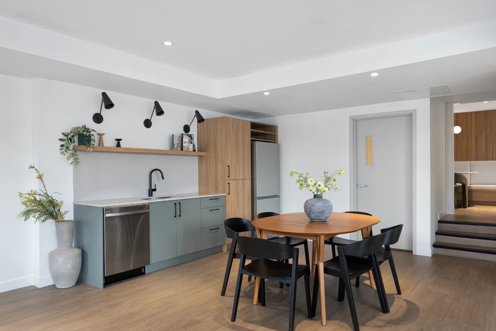 Hartley – A contemporary communal kitchen and dining area features sage green base cabinets, a stainless steel dishwasher and refrigerator, and a round wooden dining table surrounded by black chairs. White walls are accented by black wall-mounted sconces, open wood shelving, and steps leading to an adjacent lounge area with wood-paneled walls.