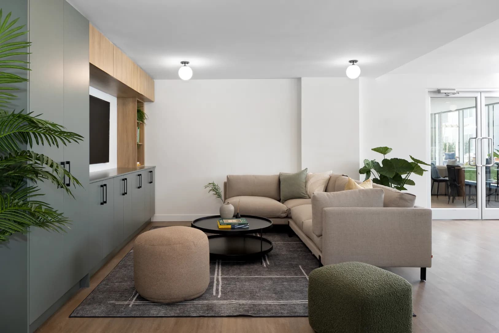 Hartley – A modern indoor lounge area features a beige sectional sofa, rounded ottomans, and a round black coffee table set on a patterned area rug. Built-in sage green cabinetry with light wood upper shelving houses a wall-mounted TV, while glass doors in the background open to an outdoor patio.