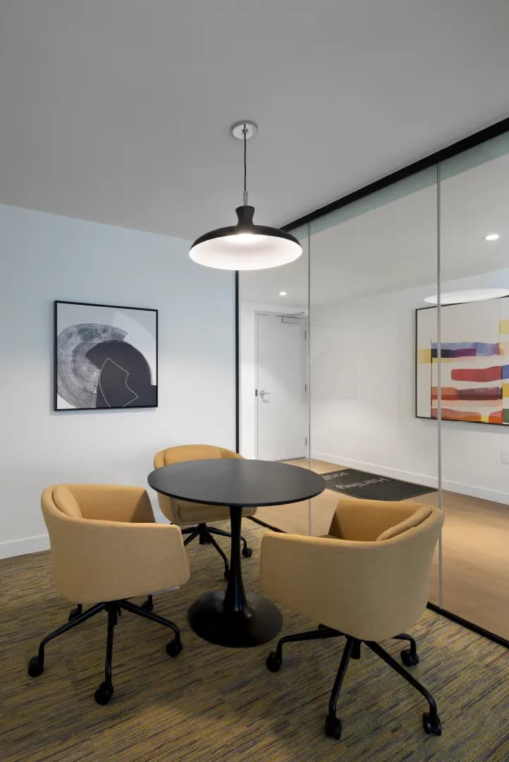 Hartley – A modern co-working or meeting room featuring a small round black pedestal table surrounded by three mustard-yellow wheeled armchairs on patterned carpeting. The space is illuminated by a black pendant light fixture and separated from an adjoining hallway by full-height glass partition walls adorned with abstract wall art.