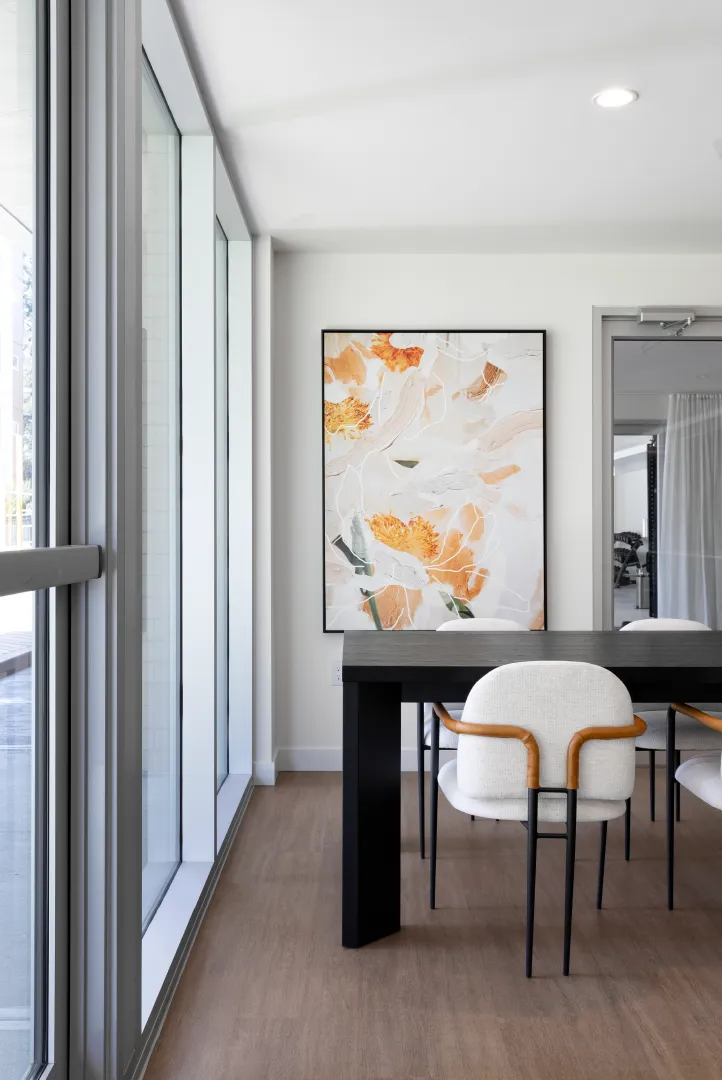 Hartley – A bright communal dining or meeting room featuring a long black table surrounded by modern white upholstered chairs with warm-toned arm accents. Floor-to-ceiling windows line the left wall, illuminating light wood-style flooring and a large abstract floral artwork hung on the neutral white back wall.