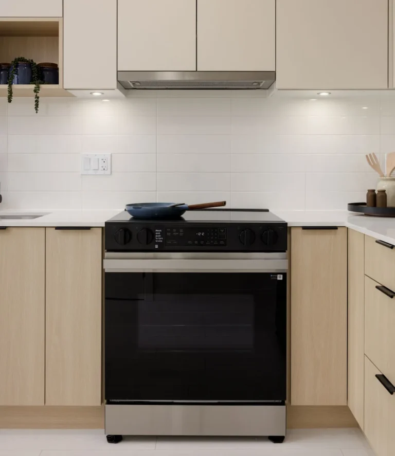Hartley – A clean, modern kitchen vignette focused on a stainless steel and black glass slide-in range set between light wood flat-panel cabinets and white countertops. The space features a glossy white subway tile backsplash, under-cabinet lighting, a built-in range hood, and minimal decorative staging like a blue frying pan and small plant.