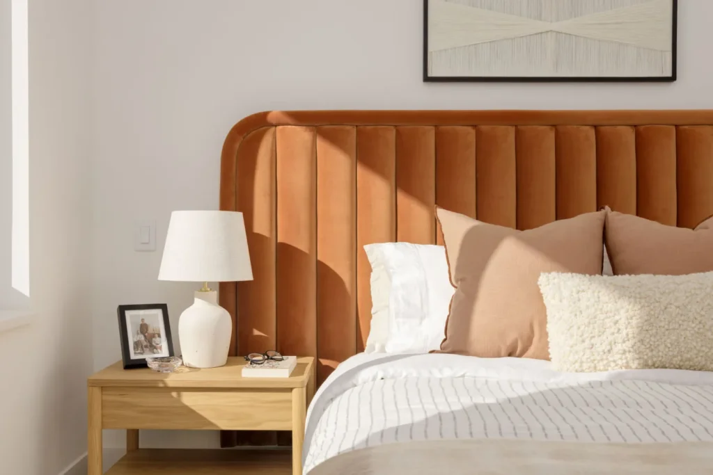 Hartley – A cozy bedroom detail featuring a bed with an ochre velvet, vertically tufted headboard dressed in layered neutral pillows and striped linens. Next to the bed, a light wood nightstand holds a white ceramic lamp and decorative items, bathed in bright, warm natural sunlight.