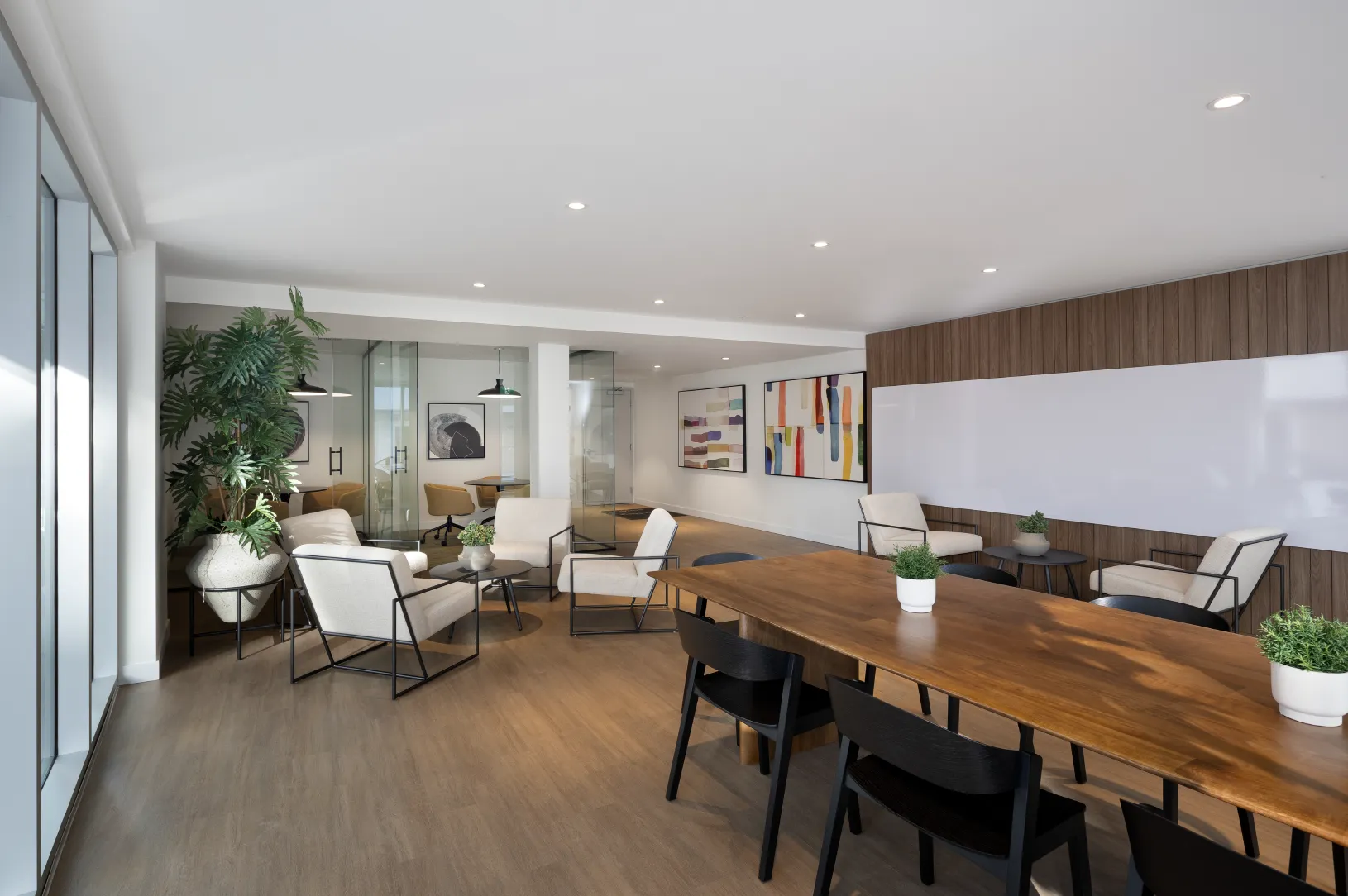 Hartley – A modern co-working and lounge amenity space with light wood flooring, white walls, and large floor-to-ceiling windows. It features a long wooden conference table with black chairs, several white accent armchairs arranged into seating vignettes, and glass-enclosed meeting rooms in the background.