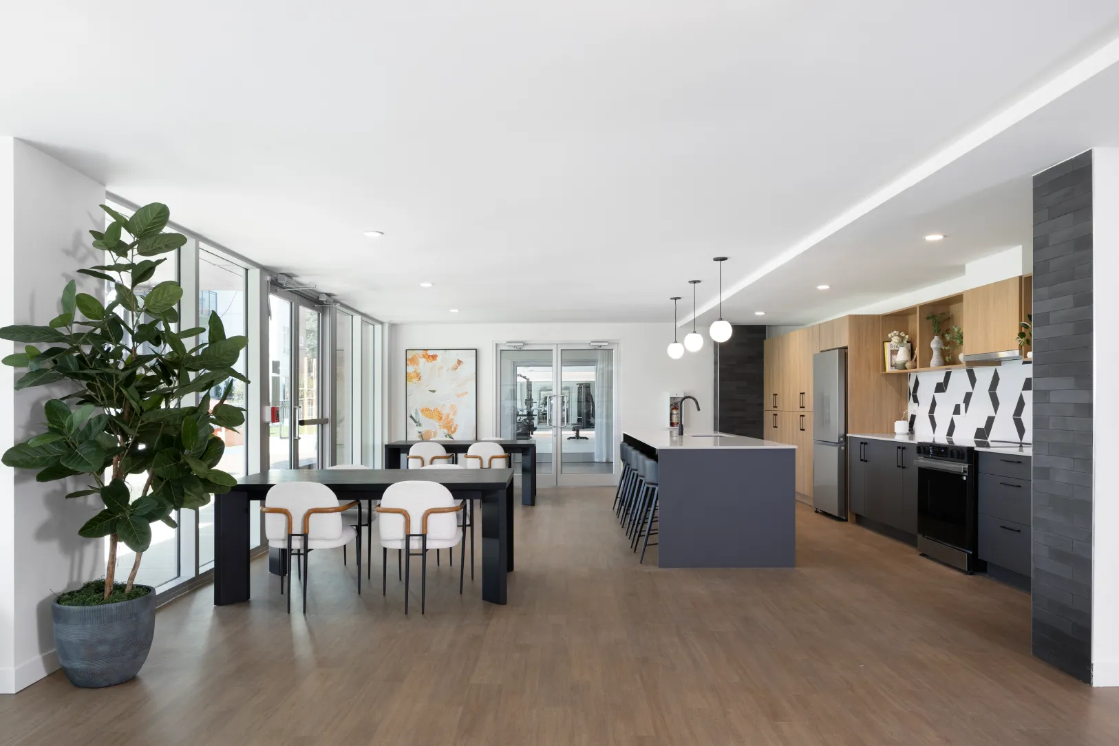 Hartley – A bright, open community clubhouse or resident lounge featuring a full kitchen with charcoal gray cabinetry, a patterned tile backsplash, and a central island with barstools. Large glass doors and windows line one wall, letting in ample natural light across wood-look flooring, multiple dining tables, and a potted tree, with a glass door in the back leading to a fitness room.