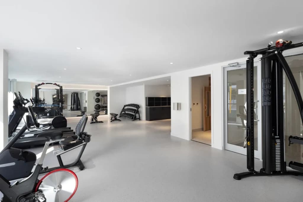 Hartley – A bright, spacious indoor fitness center featuring smooth light grey flooring, white walls, and recessed ceiling lighting. Various exercise machines are neatly spaced throughout, including stationary bikes, treadmills, a cable workout station, and free weights beside a full-length wall mirror.
