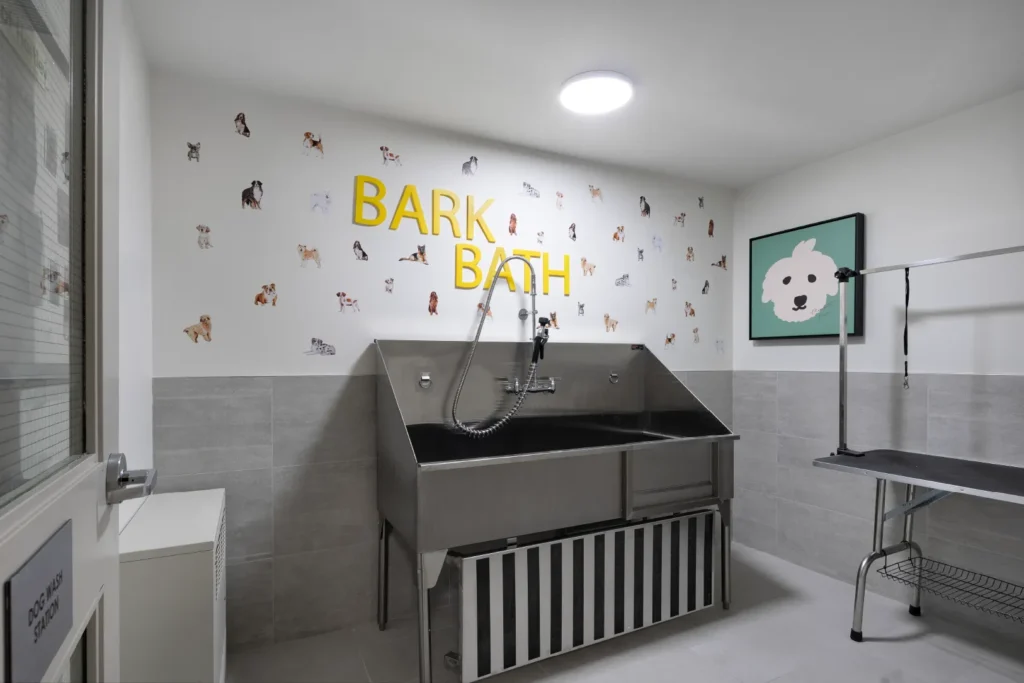 Hartley – A pet washing and grooming amenity room featuring a commercial-grade stainless steel dog washing tub and an adjustable grooming table. The space has light grey tiled lower walls, white upper walls decorated with dog stickers and yellow 'BARK BATH' lettering, and bright overhead lighting.