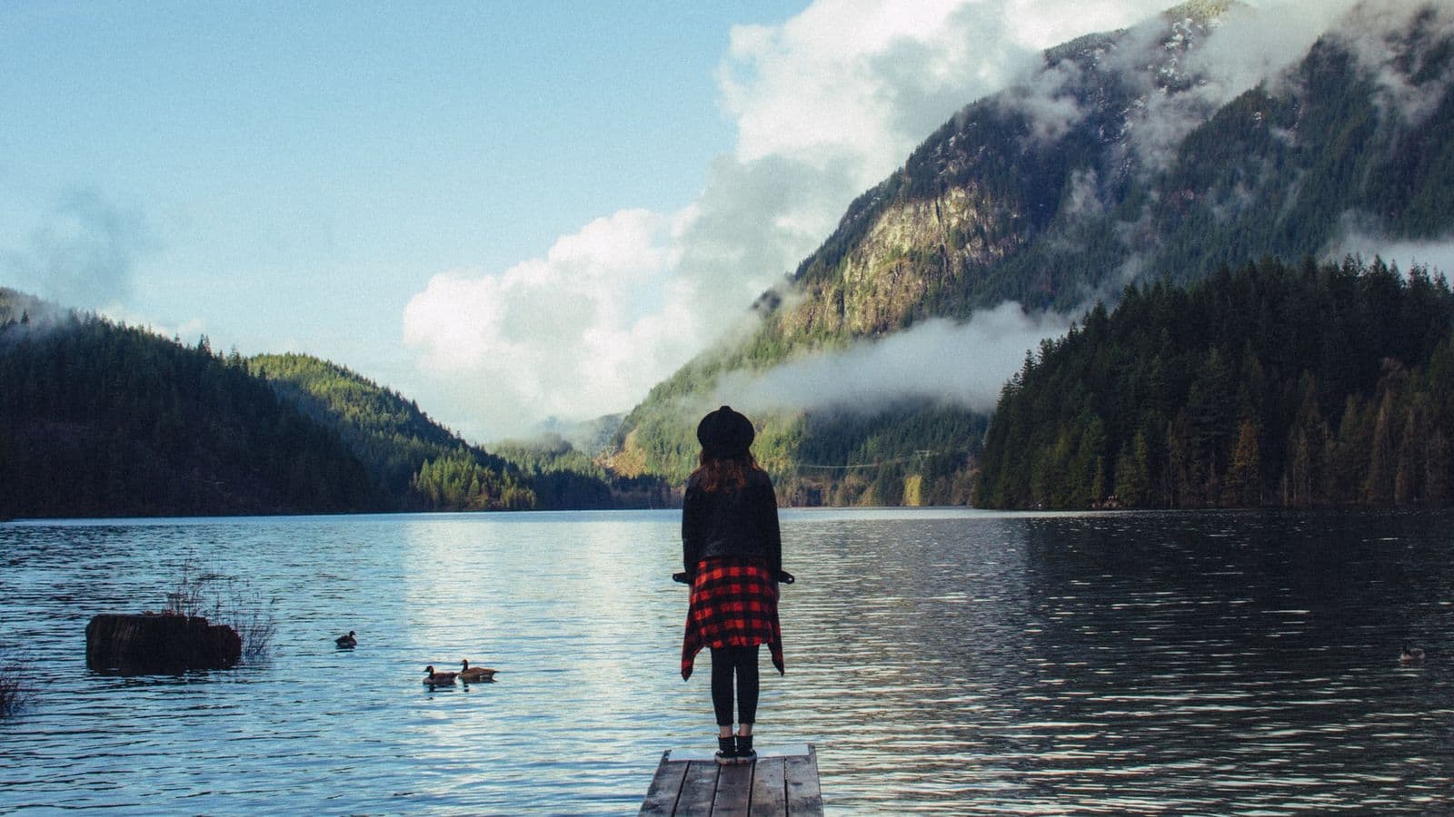 Mary Annes Place – A generic lifestyle photo of a person standing on a wooden dock looking out at a lake surrounded by forested mountains; not clearly tied to the building.