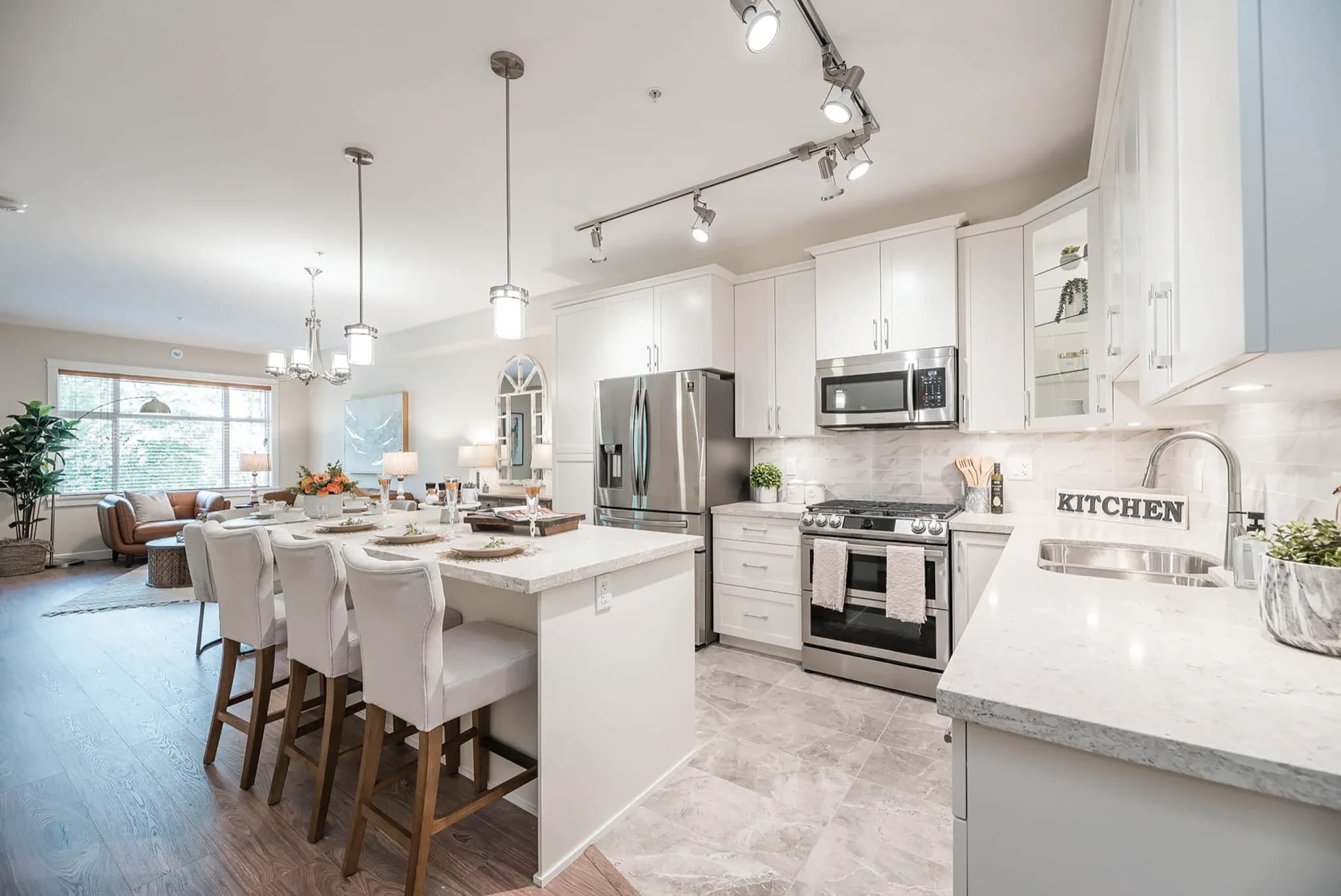 Ledgeview II – A bright, open-concept kitchen and living area featuring white shaker-style cabinetry, quartz countertops, stainless steel appliances, and a central island with bar stool seating. Modern track lighting and pendant lamps illuminate the space, which flows past a set dining area into a cozy living room with large windows.