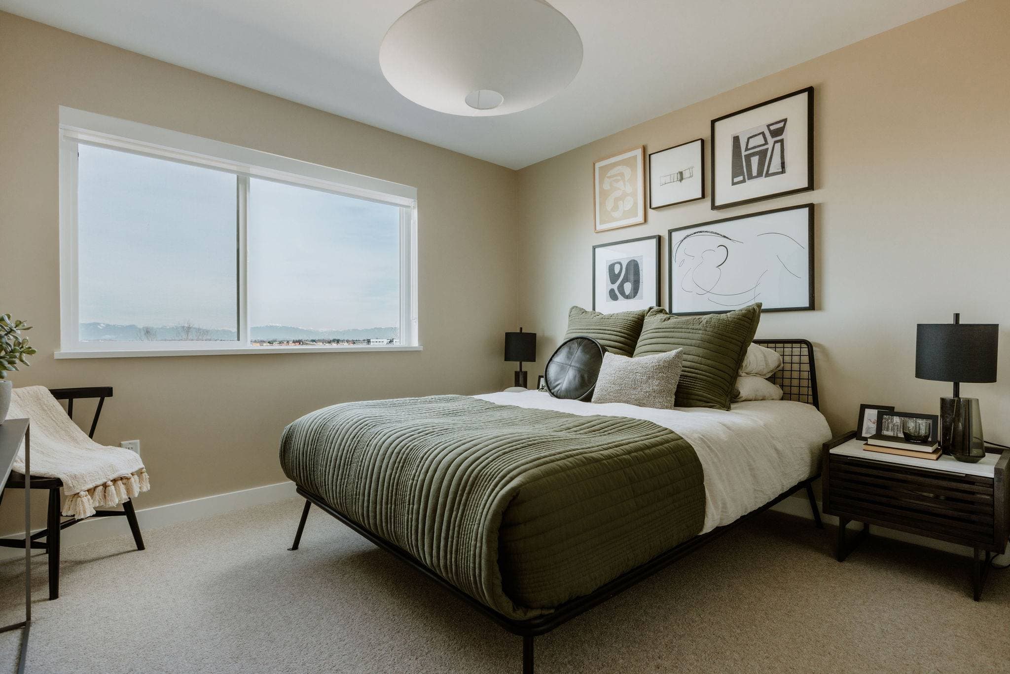Boardwalk 7 – A well-lit bedroom features a bed styled with textured olive green and white bedding, framed abstract art hanging on beige walls, and light-colored carpeting. A large window brings in daylight and offers distant views of mountains on the horizon.