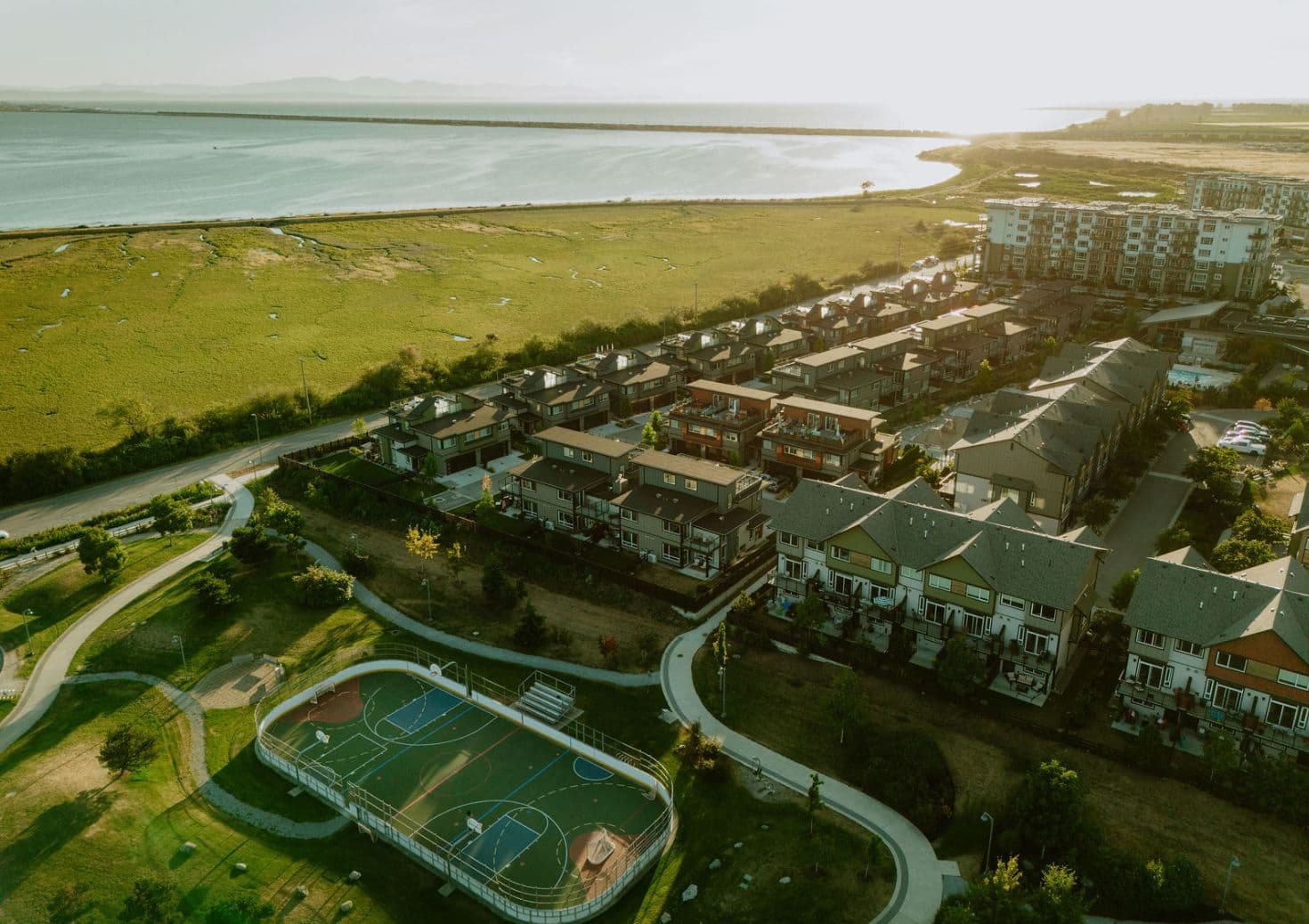 Boardwalk 7 – Aerial drone photo of a waterfront townhome community with rows of pitched-roof homes, an outdoor sport court, walking paths, and open marshland and water beyond.