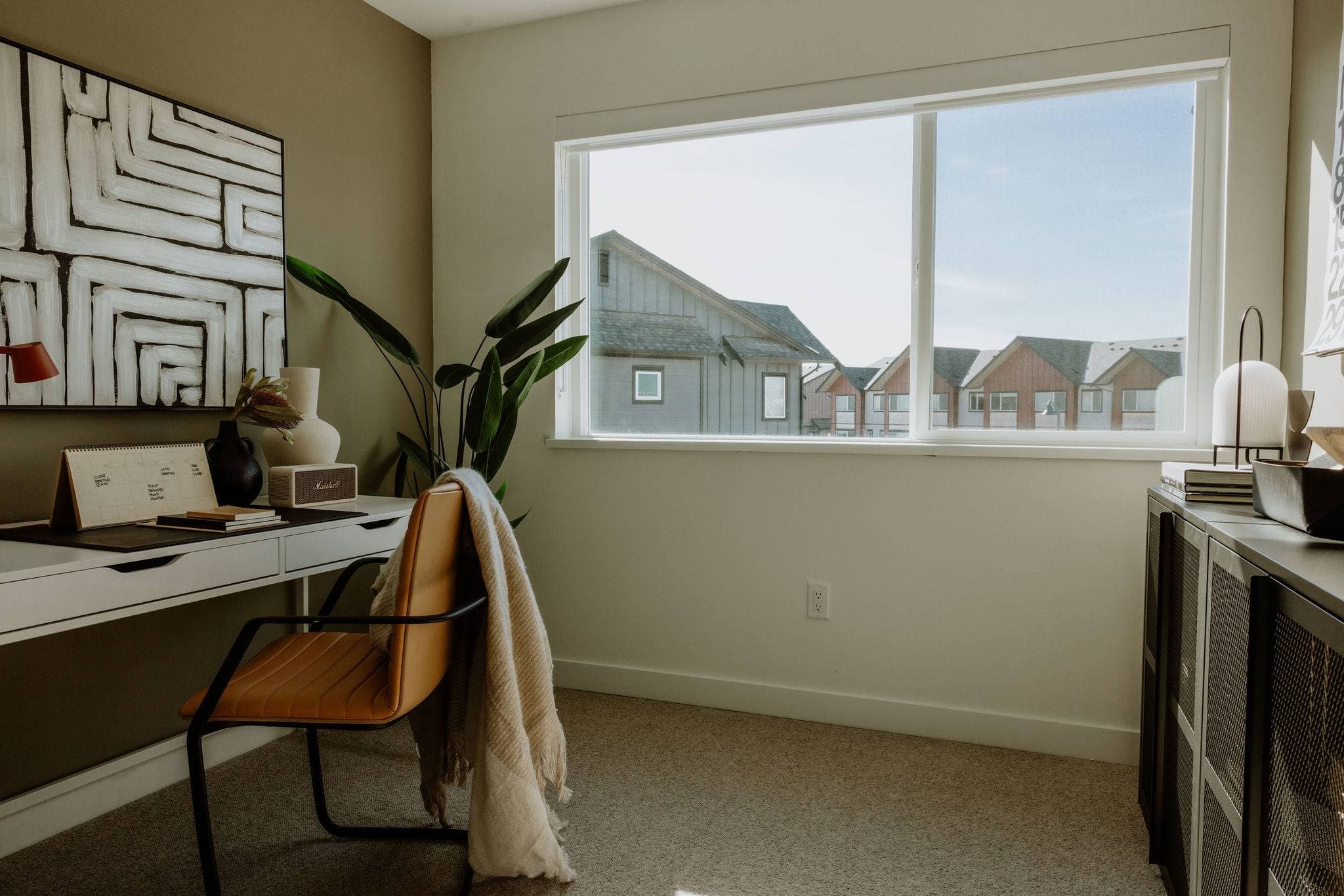 Boardwalk 7 – A home office setup featuring a white desk with a leather chair, abstract wall art, and an indoor potted plant. A large sliding window brings in bright daylight and offers a view of neighboring townhomes under a clear sky.