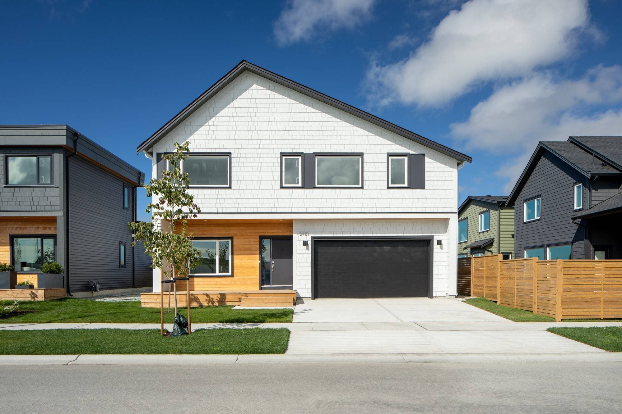 Boardwalk 7 – A contemporary two-story single-family home features white shake siding, a pitched black roofline, warm wood horizontal accents around the covered front porch, and a sleek dark garage door with a concrete driveway. Taken from street level on a bright sunny day under a blue sky with light clouds, the house includes a neatly manicured front lawn and is bordered by wooden fences and neighboring moder