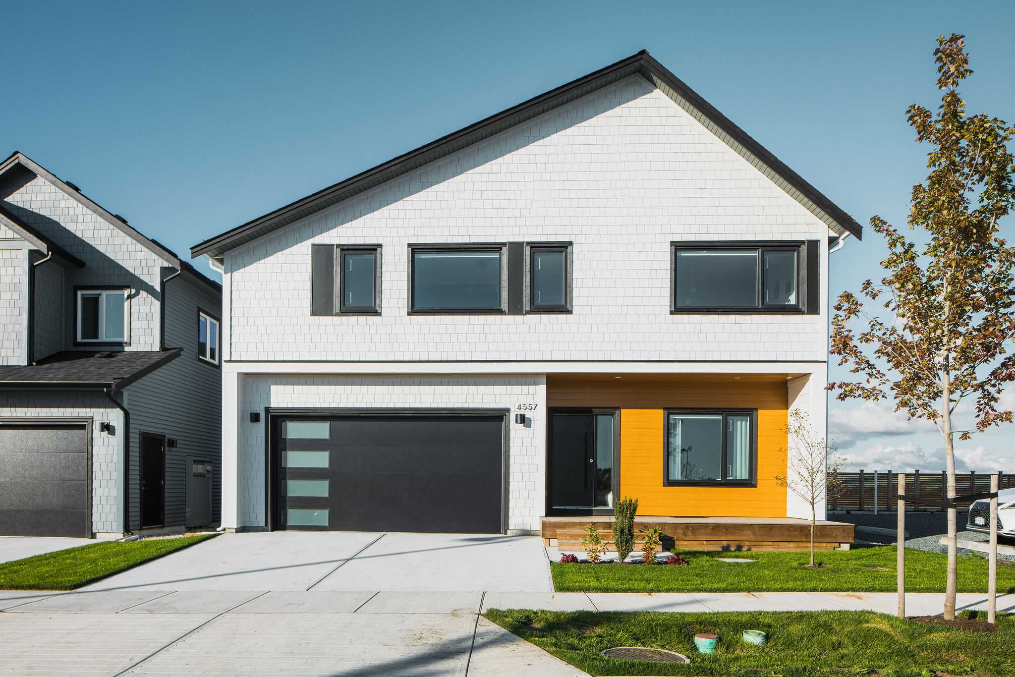 Boardwalk 7 – A contemporary two-story house captured from the driveway on a sunny day, featuring white shingle siding, dark-trimmed windows, a modern black garage door, and a vibrant warm-wood accent wall framing the front entrance. A concrete driveway and manicured green lawn sit in the foreground, with an adjacent grey home partially visible on the left.
