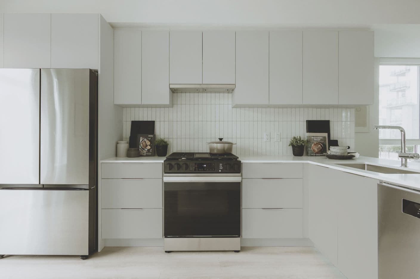 Boardwalk 7 – A clean, minimalist kitchen featuring flat-panel light gray cabinetry, stainless steel appliances, and a vertical white tile backsplash. Natural daylight fills the space, which includes an undermount sink, integrated gas range, and subtle countertop styling with cookbooks and small potted plants.