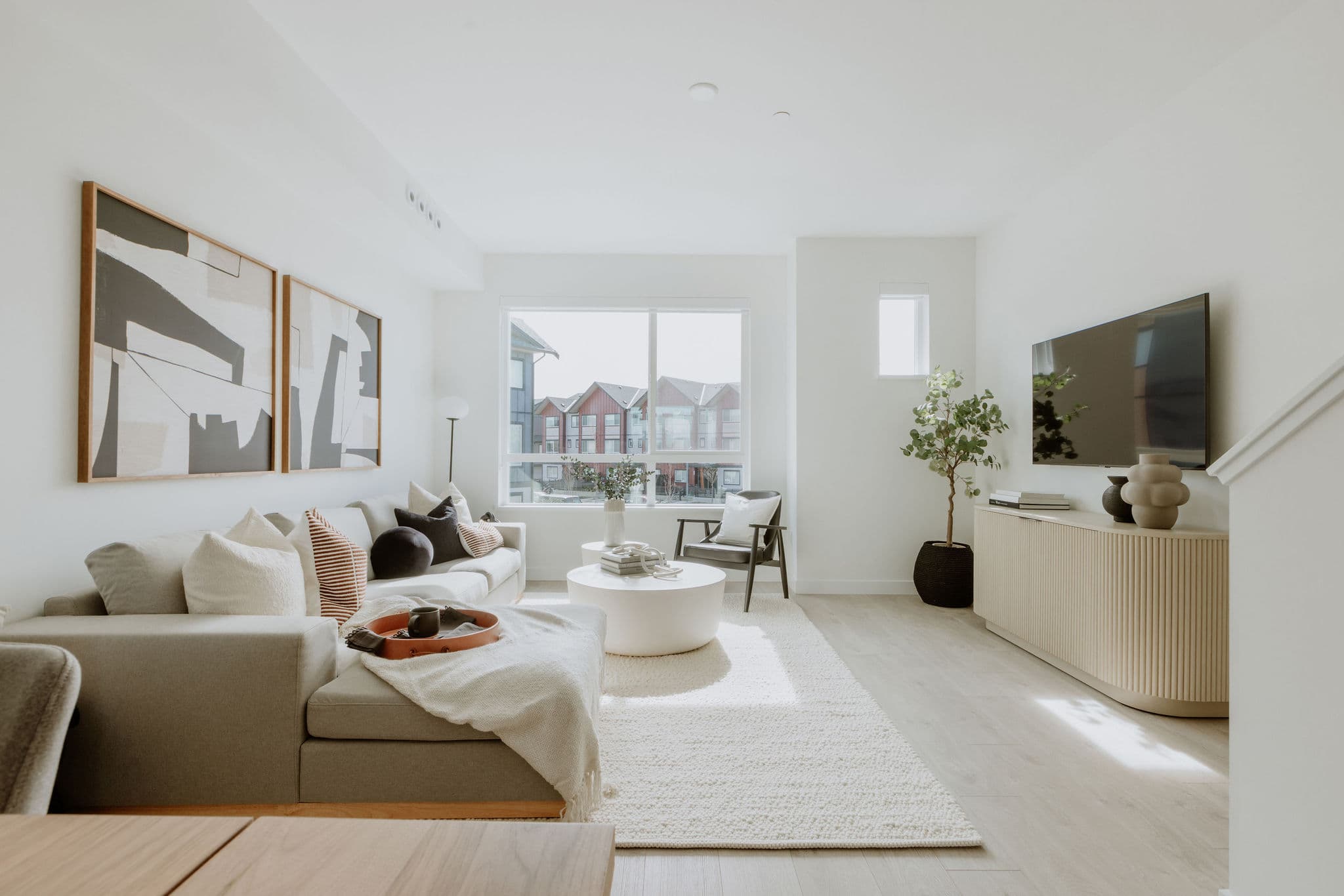 Boardwalk 7 – A bright and airy modern living room features a light grey sectional sofa adorned with pillows, a round white coffee table, and light wood flooring topped with a cream rug. Sunlight streams through a large window overlooking adjacent townhomes, highlighting crisp white walls, abstract framed artwork, a fluted TV console, and a potted indoor tree.