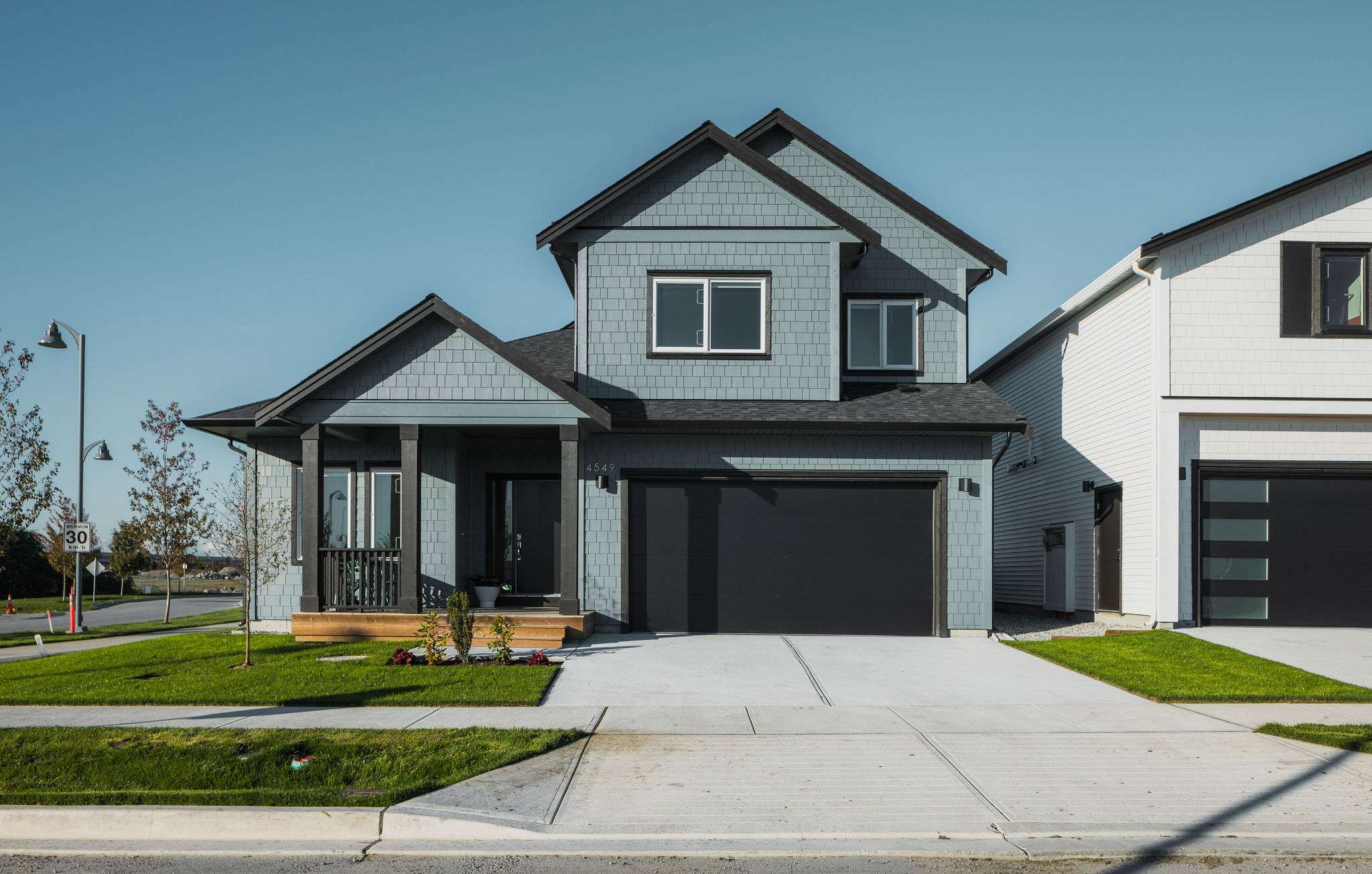 Boardwalk 7 – A modern two-story single-family home featuring dusty blue shake siding, black trim, and a black double-car garage door. A concrete driveway and manicured front lawn lead up to a covered front porch under a clear blue daytime sky.