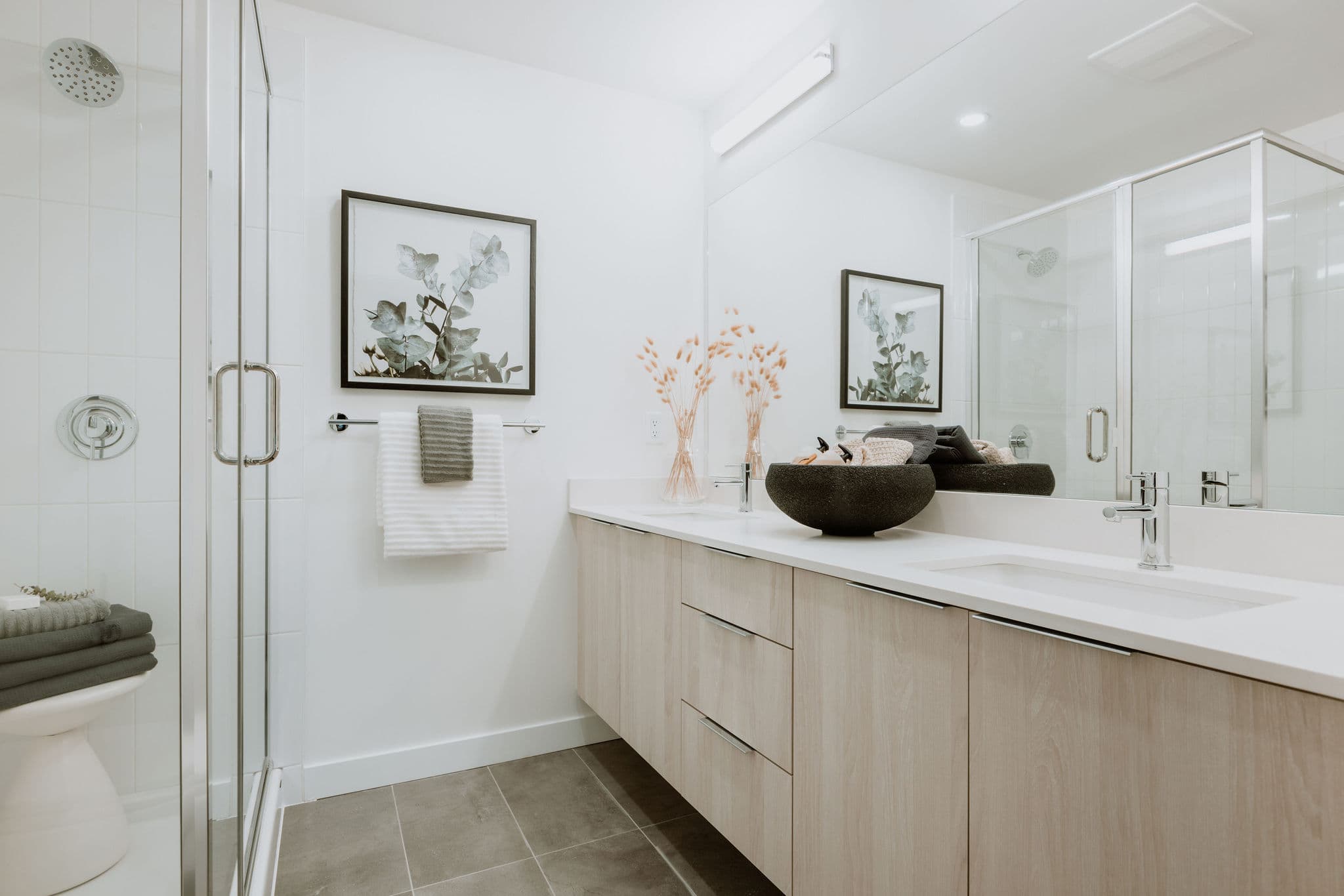 Boardwalk 7 – A modern, bright bathroom featuring a floating light wood double vanity with white countertops and polished chrome fixtures. A glass-enclosed walk-in shower is adjacent to the vanity, complemented by clean white walls, grey floor tiles, and a large unframed mirror.