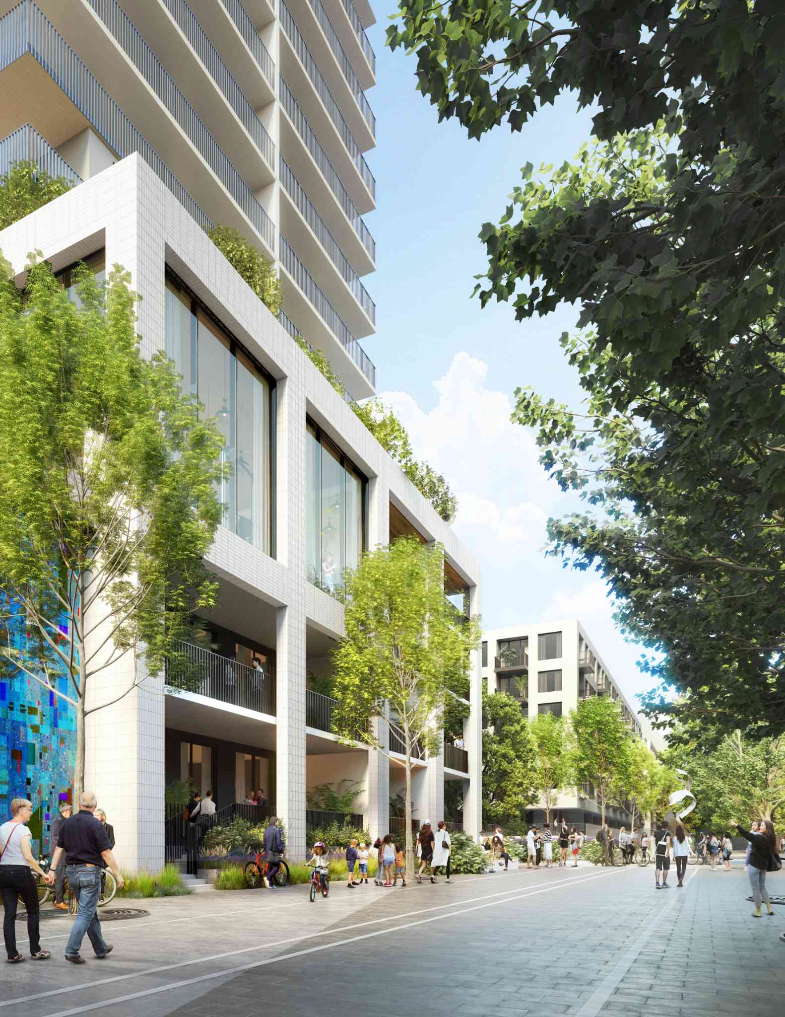 Lansdowne District – A ground-level streetscape view showcasing a modern residential building with a white tiled facade, double-height windows, and balcony terraces framed by mature green trees. Pedestrians, cyclists, and families populate the wide, paved walkway on a bright, sunny day.