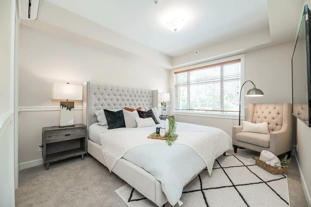 Walnut Park – Bedroom staged with a grey tufted upholstered bed, nightstands with lamps, an armchair, and a large window.