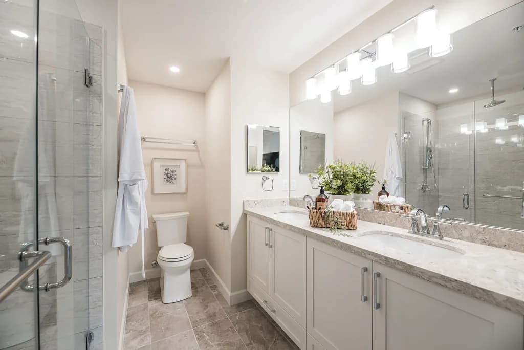 Walnut Park – Bathroom with double sink vanity, glass shower with rainhead, framed art, and a robe hanging by the toilet.