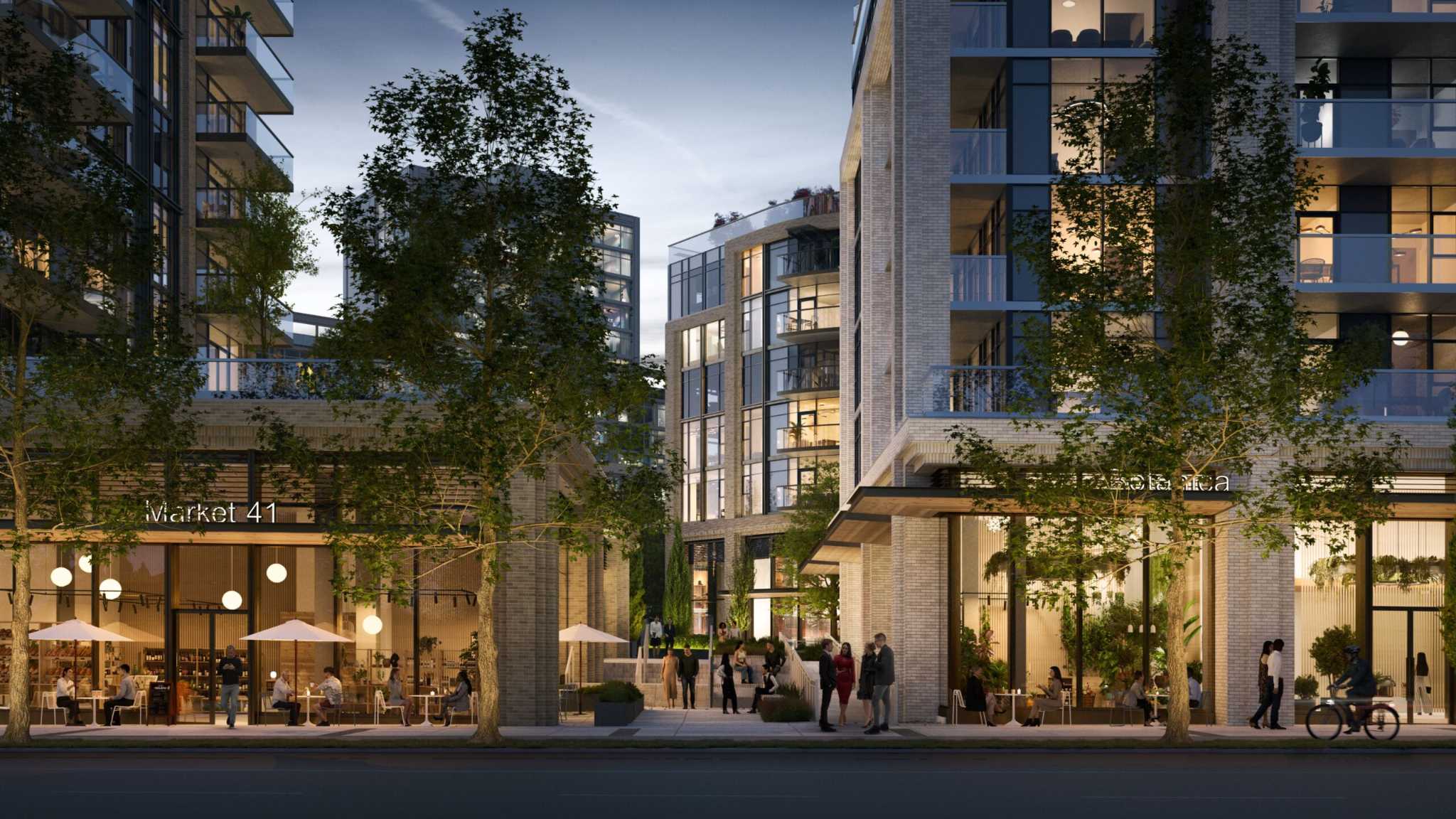 Mayfair West – A twilight street-level view of contemporary multi-story brick and glass residential buildings featuring bustling ground-floor retail and cafes. Pedestrians walk along the sidewalk and through a landscaped courtyard between the brightly illuminated storefronts.