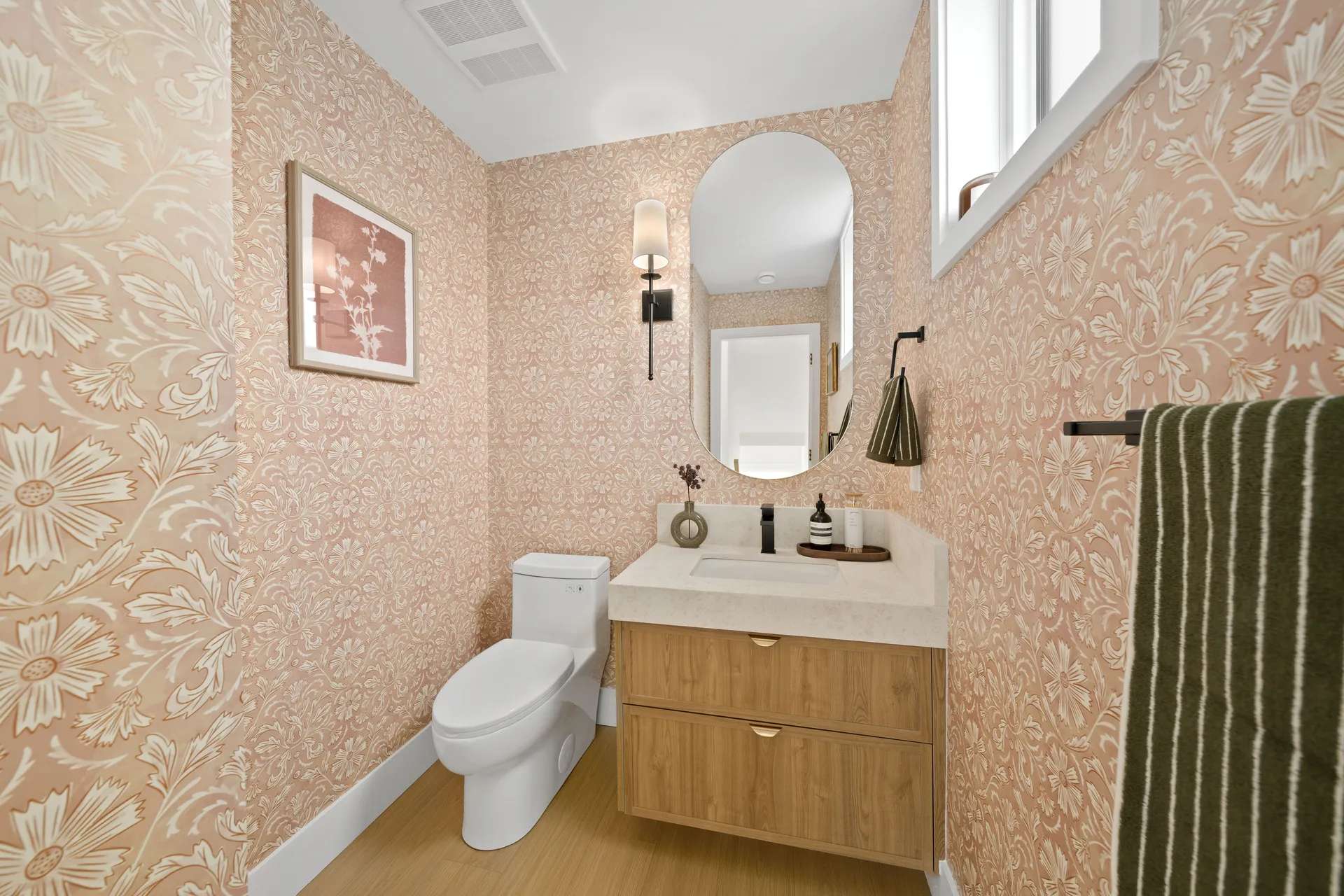 Madison Townhomes – Powder room with pink floral wallpaper, an oval mirror, a wood vanity, and a white toilet.