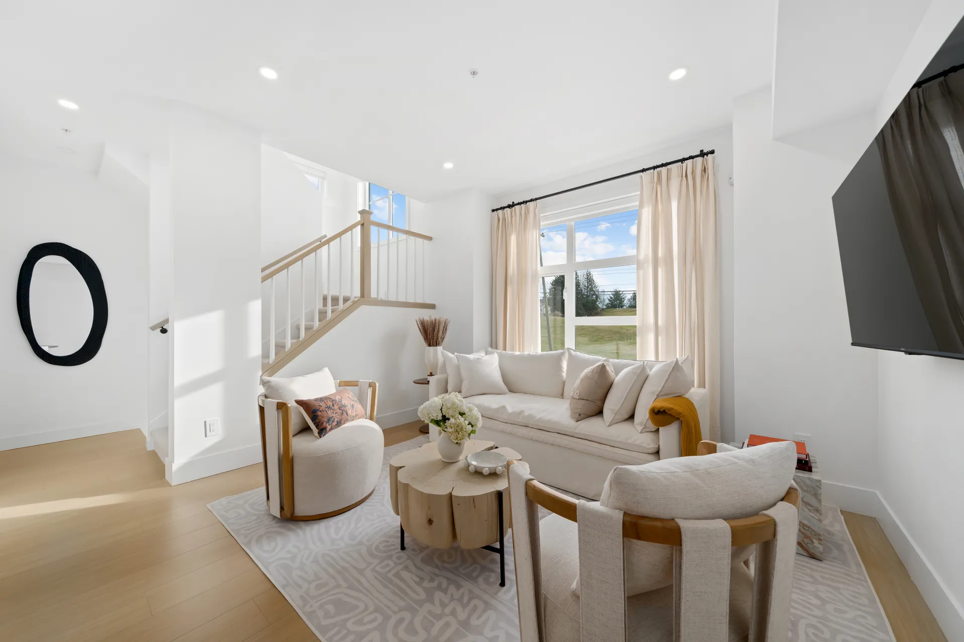 Madison Townhomes – Living room with a cream sofa and armchairs, a round wood coffee table, a wall-mounted TV, and a staircase to an upper floor.