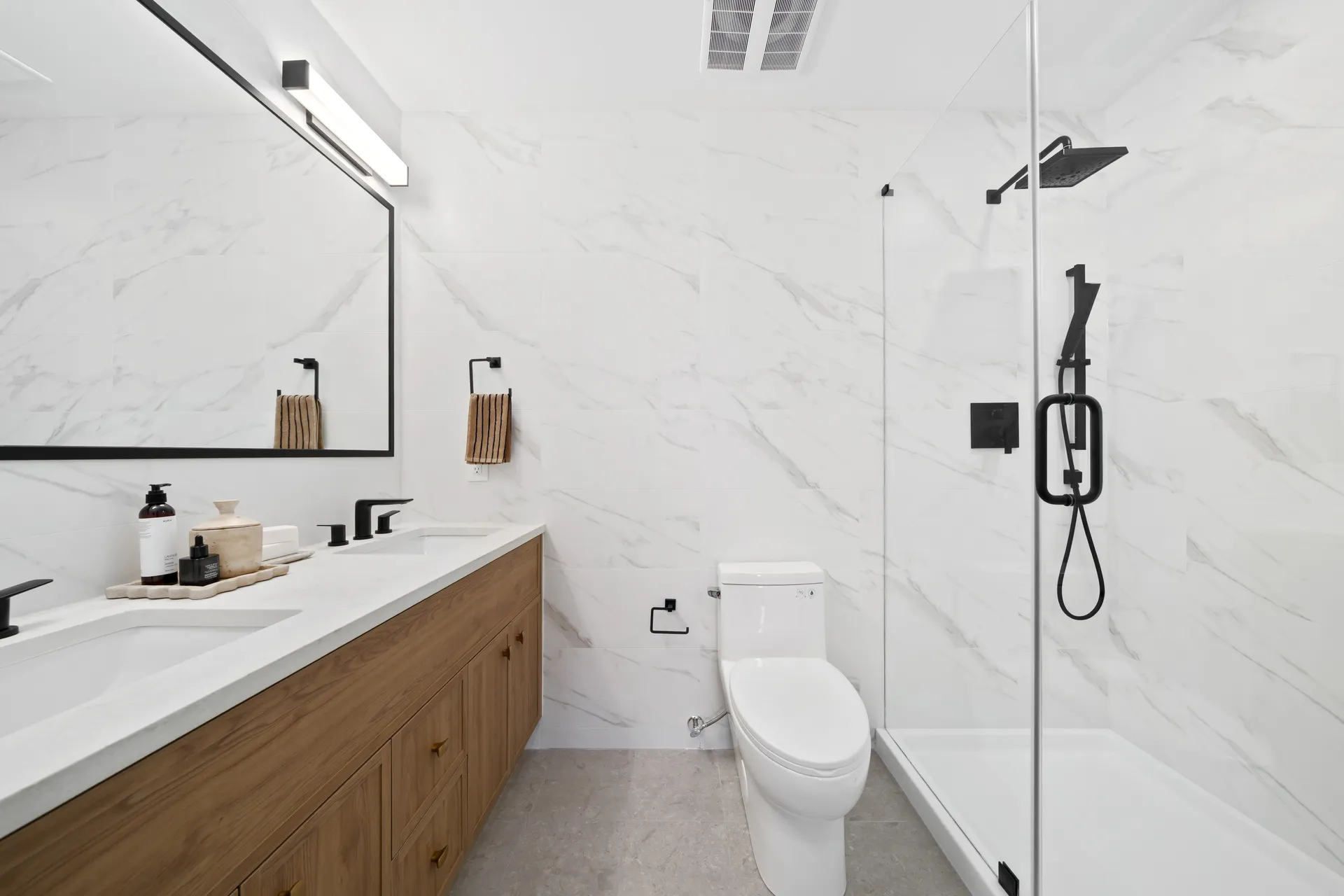 Madison Townhomes – Bathroom with a glass-enclosed shower, marble-look tile, a white toilet, and a wood vanity with double sinks.