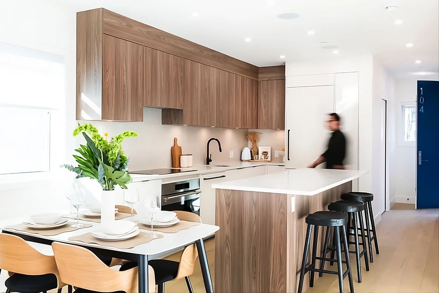 Coral – A bright, modern kitchen and dining area featuring wood-paneled upper cabinetry, a matching island with black barstools, white countertops, and an adjacent dining table set for four.
