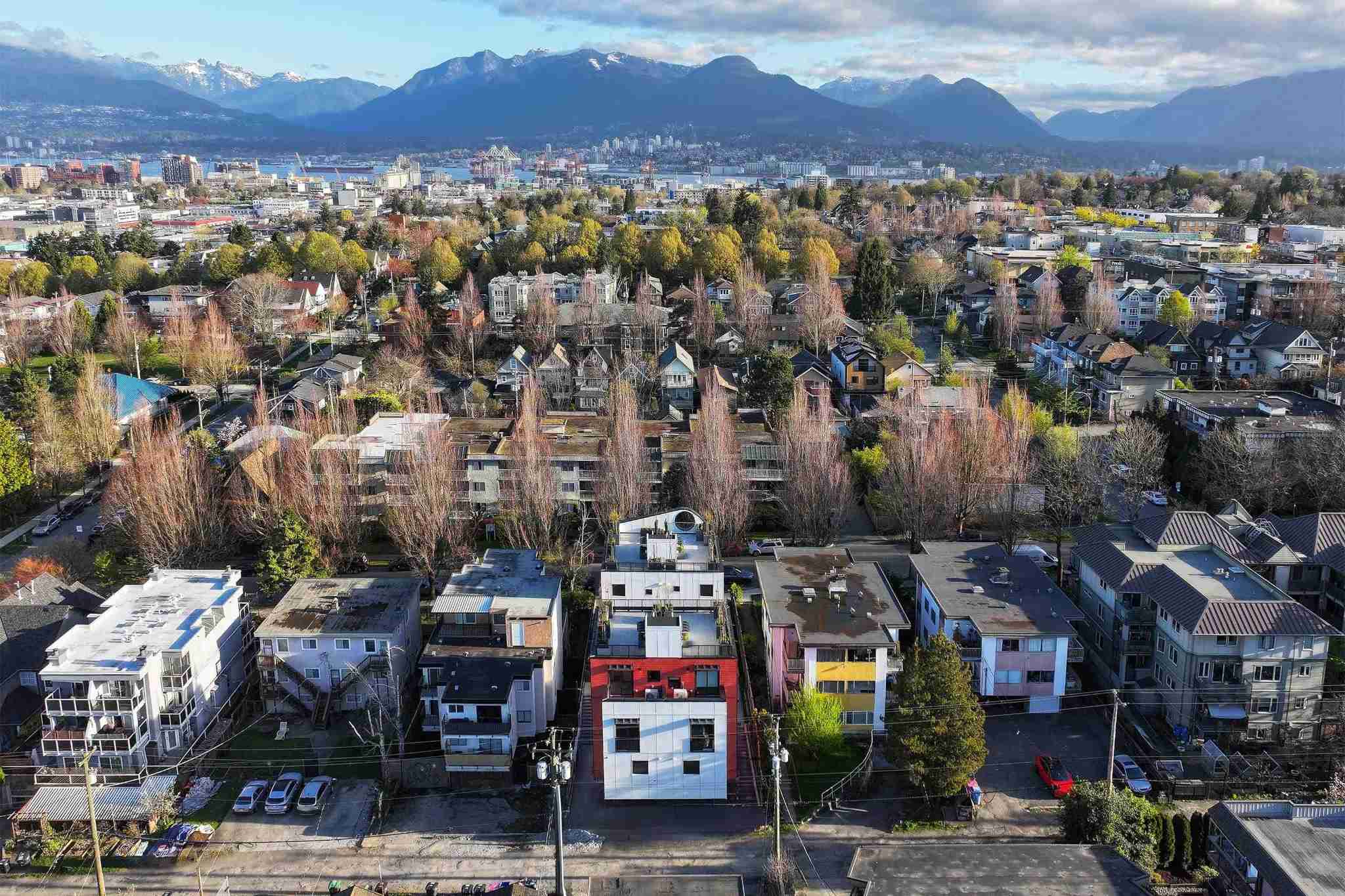 Coral – An elevated neighborhood overview features a modern white and red residential building among tree-lined streets and low-rise homes. The cityscape, port, and snow-capped mountain range stretch across the horizon beneath a partly cloudy sky.