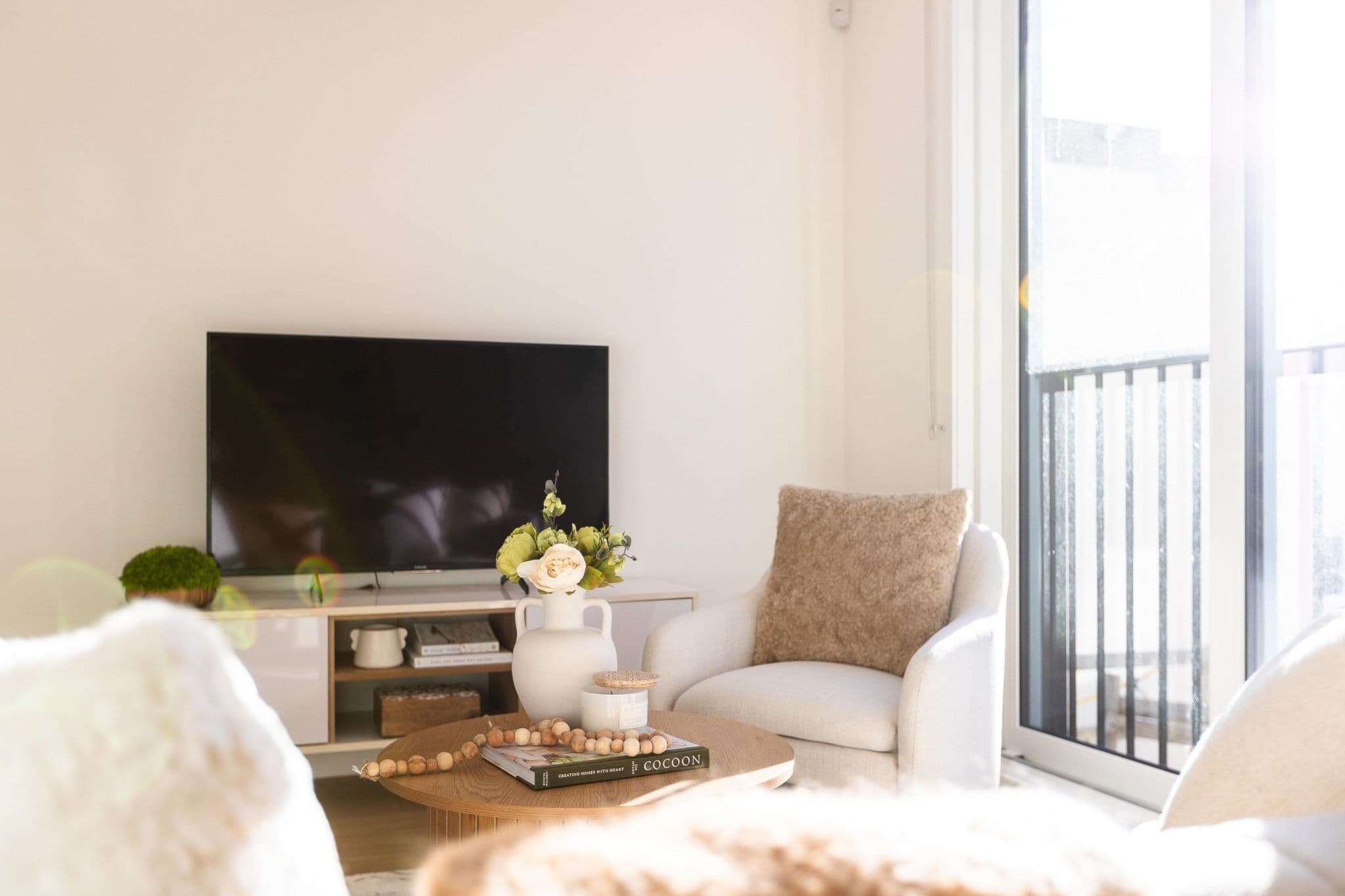 Coral – A sun-drenched modern living room with neutral decor, featuring an upholstered armchair, a round wooden coffee table, and a flat-screen TV on a low media console. Natural light floods in from a large glass door leading to a balcony.