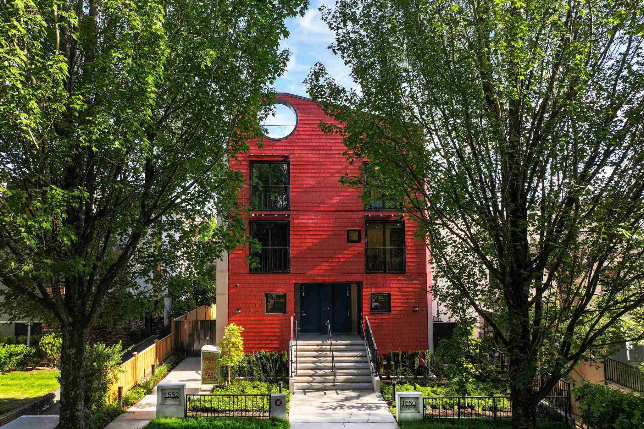 Coral – A bright red, shingle-clad contemporary residential building is framed by lush green trees under a clear blue sky. The front entrance features landscaped walkways, concrete stairs, and a distinctive round window near the roofline.