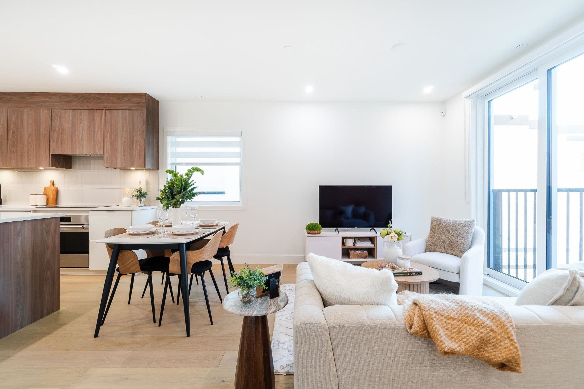 Coral – A bright open-concept living and dining area featuring light wood flooring, a four-person dining set, and an adjacent kitchen with warm wood upper cabinets. Large sliding glass doors lead to a balcony next to a contemporary seating area with a sofa, armchair, and media console.