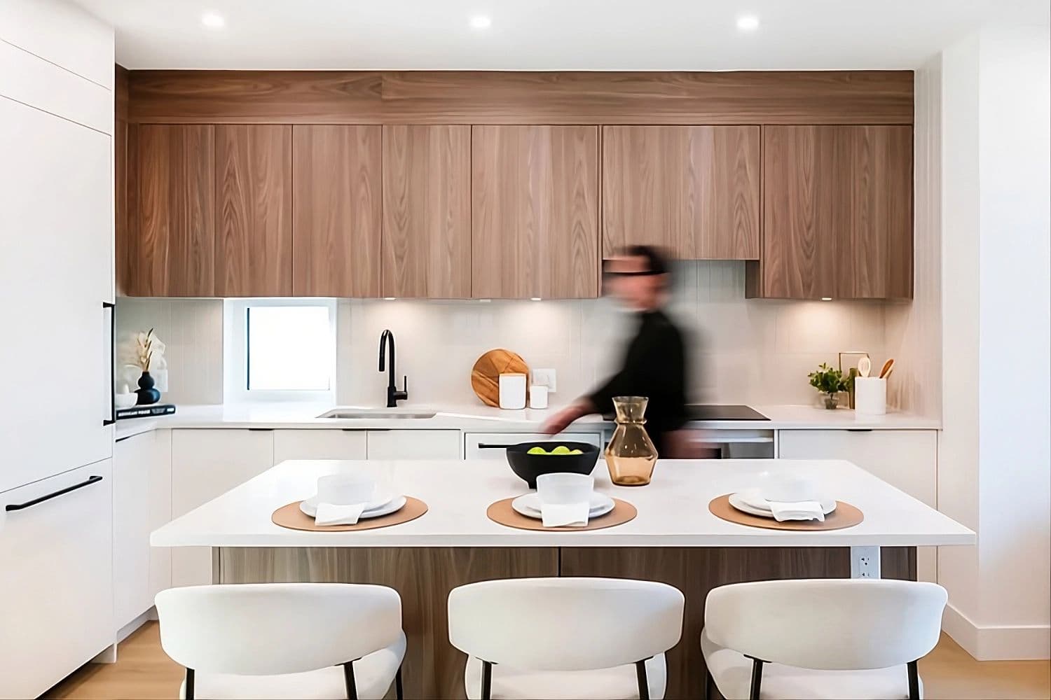 Coral – A modern kitchen showcases warm wood upper cabinetry, crisp white counters, and a central island with three barstools and place settings. A motion-blurred person stands in the bright, warmly lit minimalist space.