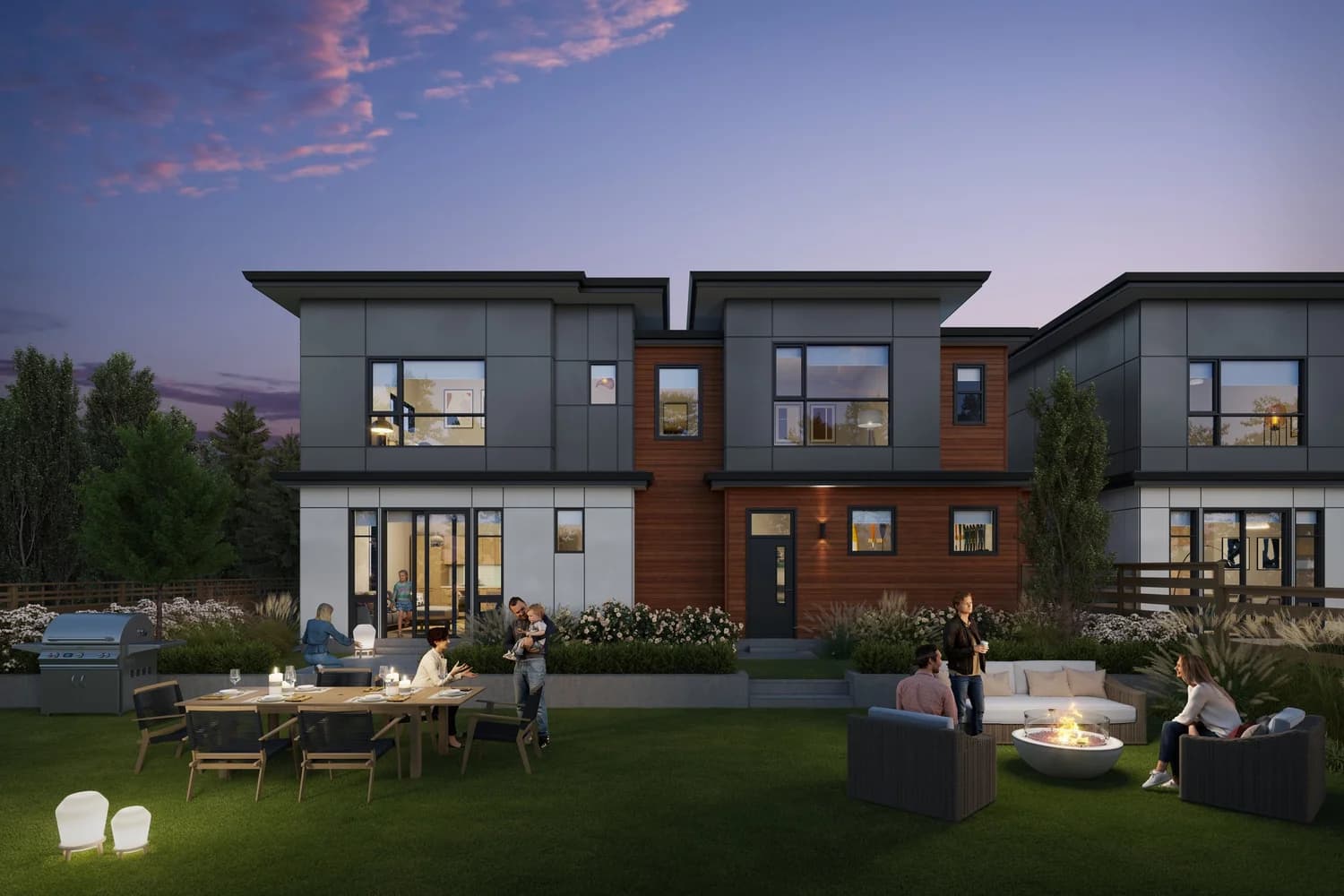 Onward – A dusk view of the backyard of modern two-story townhomes clad in dark gray panels and warm wood siding. People gather across a spacious green lawn furnished with an outdoor dining table, a barbecue grill, and an illuminated fire pit lounge area under a purple twilight sky.