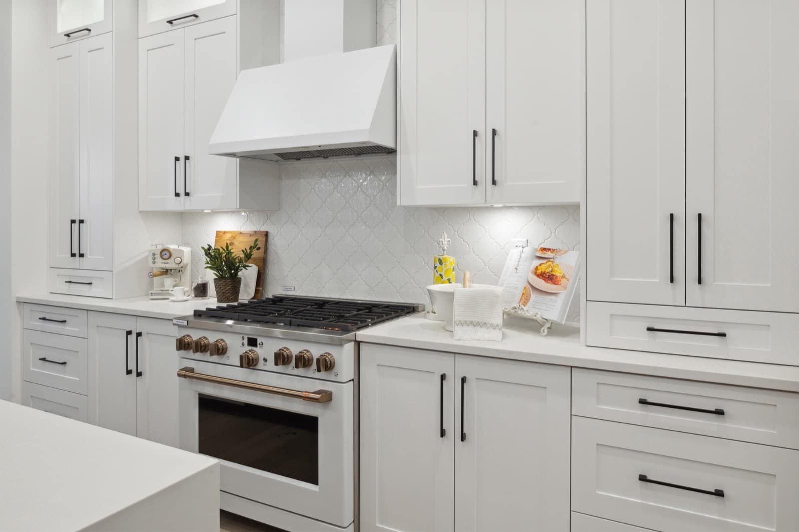 Brookswood Estates – A white kitchen with a white range hood, tile backsplash, and a coffee bar area with espresso machine and cookbook stand on the counter.